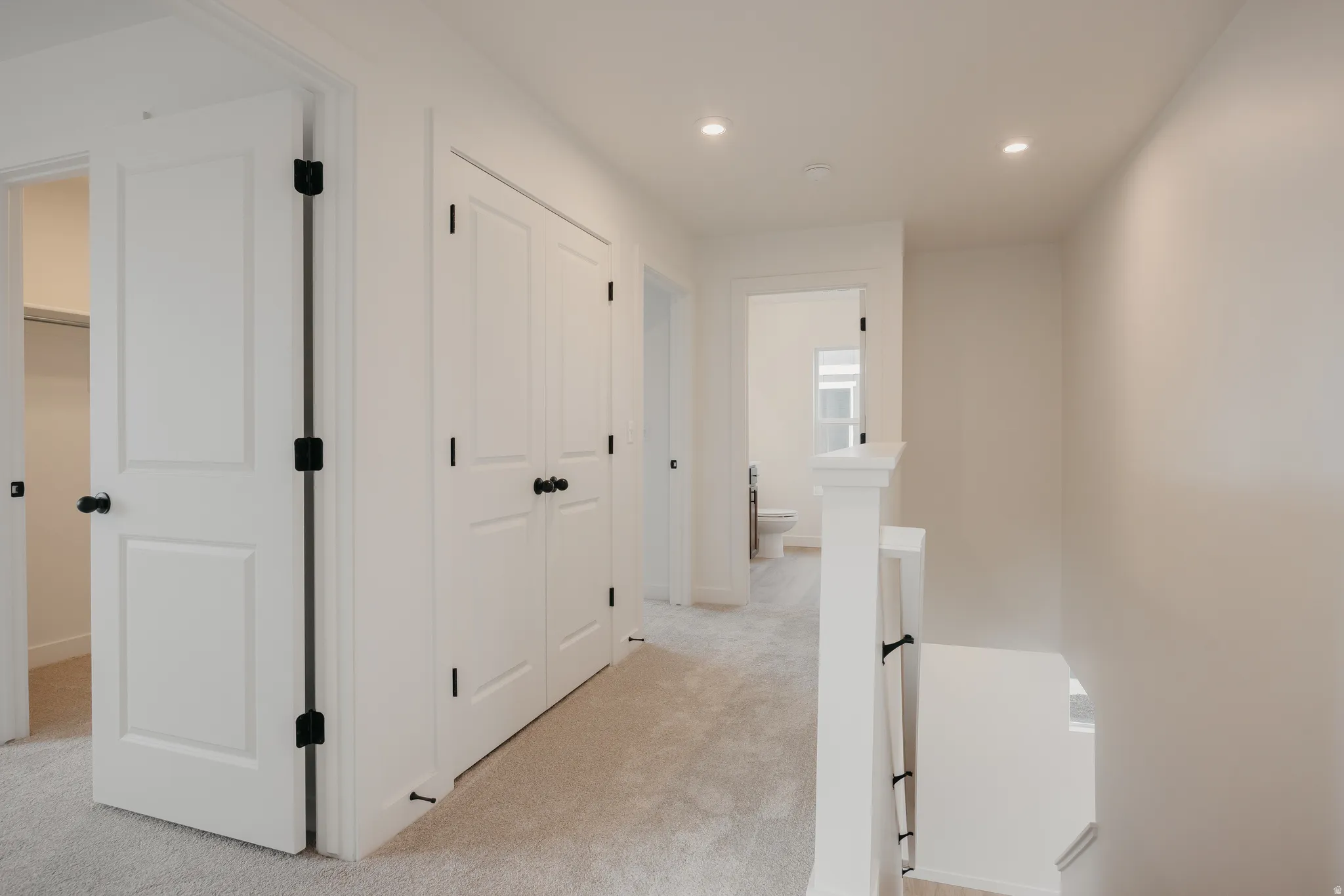 Carpeted hallway featuring bright white walls, recessed lighting, white panel doors with matte black hardware, and a white staircase with black iron balusters