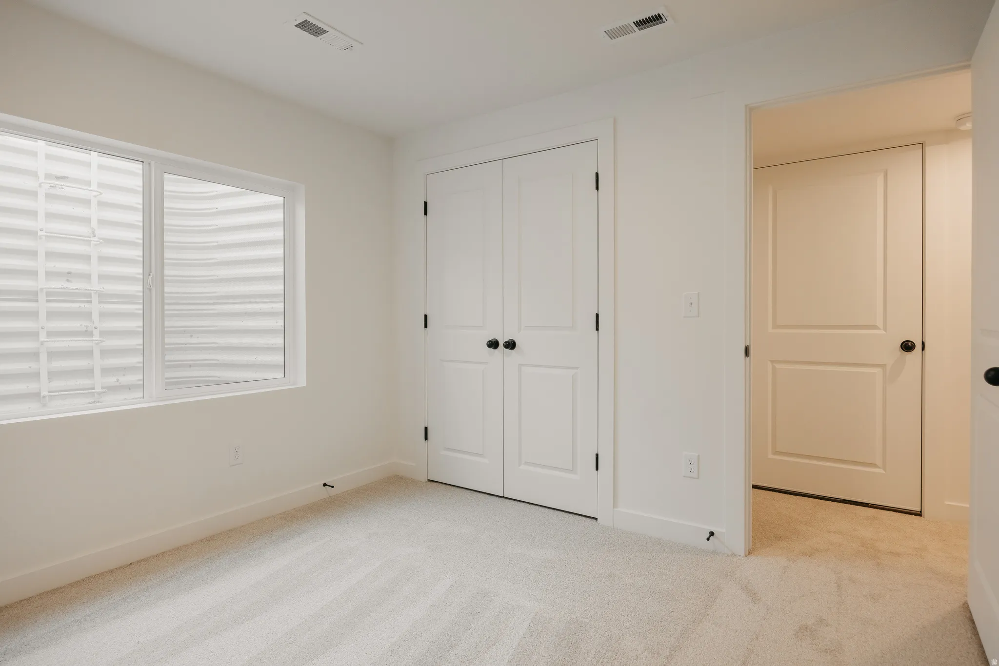 Bedroom featuring light-toned carpet, a window with a window well, white baseboards, and white two-panel doors with black hardware