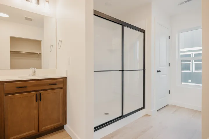 Contemporary bathroom featuring a black-framed glass shower enclosure, a wood-finish vanity with a white countertop and integrated sink, a large wall mirror, and light wood-finish flooring