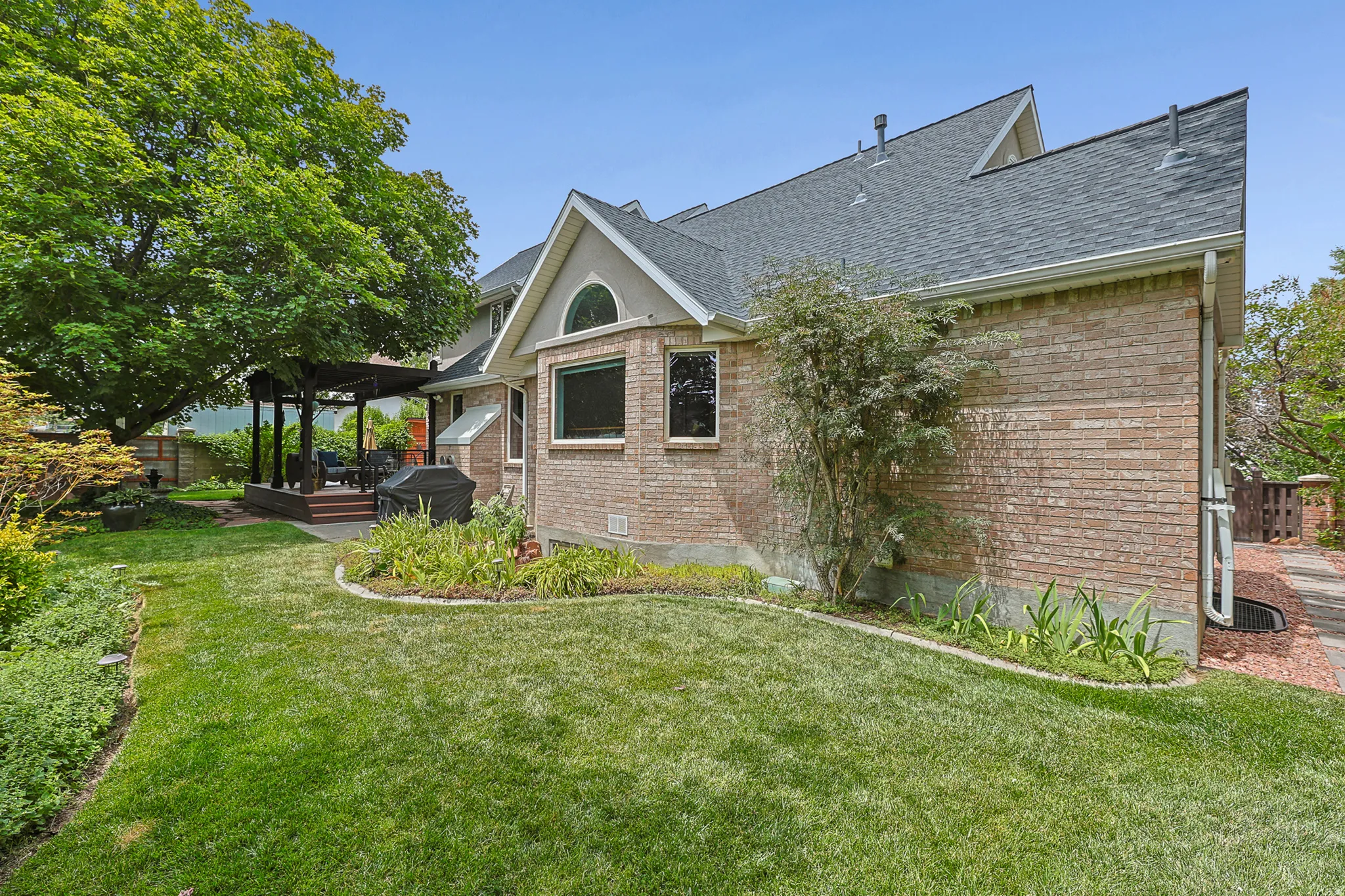 Brick exterior with a gabled roofline, featuring a covered pergola on a raised trek deck with redwood pergula, mature trees, and a manicured lawn with garden beds