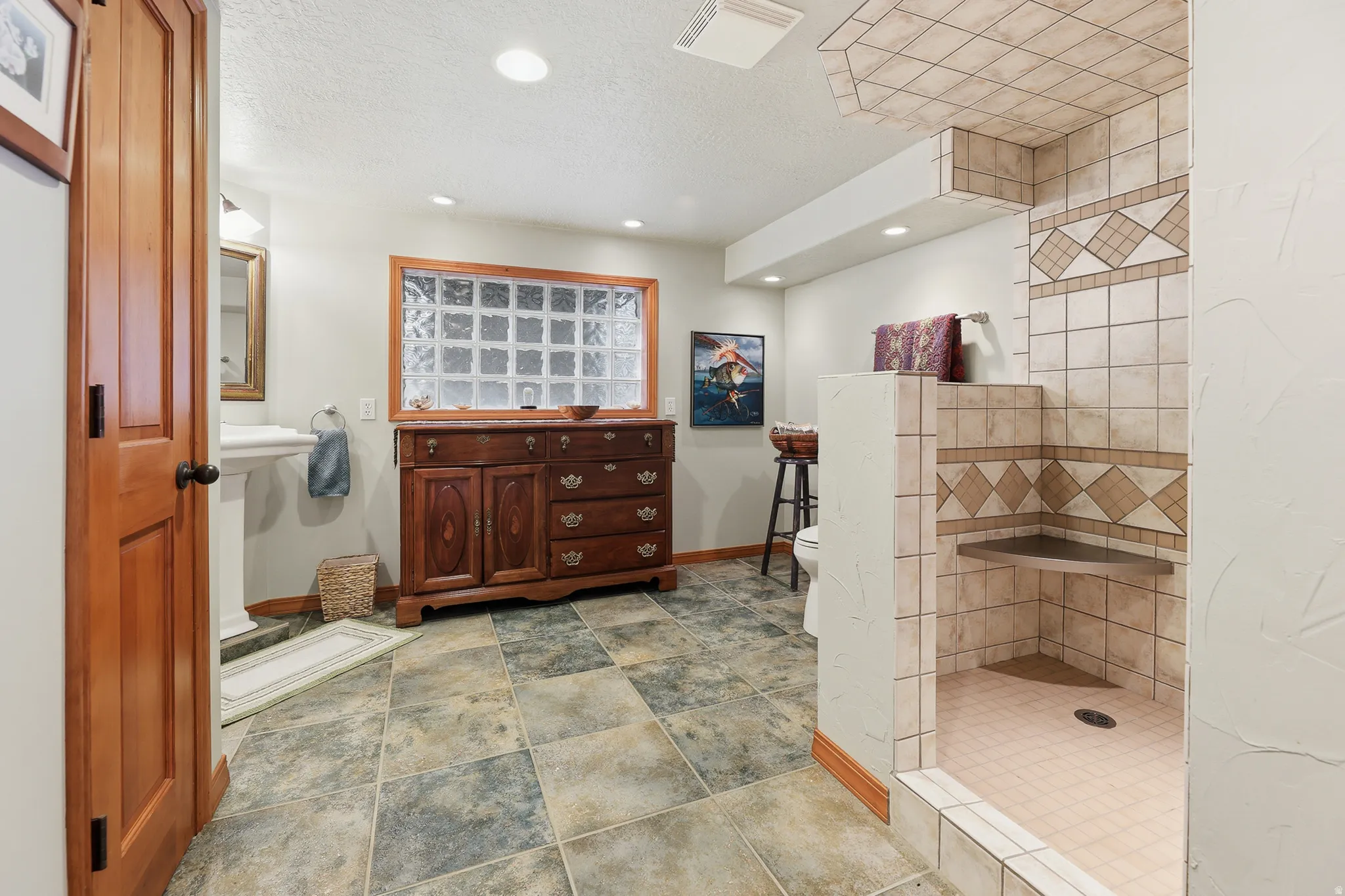 Spacious bathroom featuring an open tiled shower with a built-in bench, a glass block window, a pedestal sink, recessed lighting, and large format stone-look floor tiles