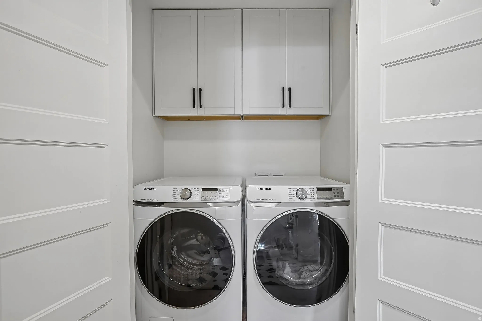 Dedicated laundry closet featuring white paneled doors, white upper cabinetry with black hardware, and a wood shelf