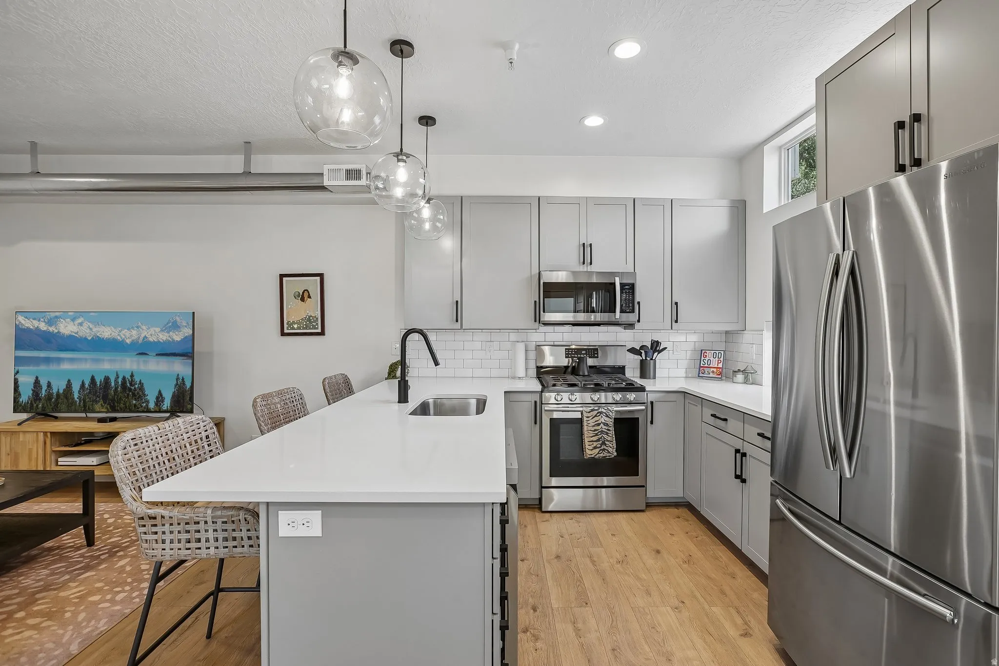 Modern kitchen featuring light gray cabinetry, stainless steel appliances, white subway tile backsplash, a large kitchen island with a white countertop and undermount sink, and wood-finish flooring