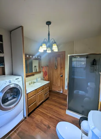 Bathroom featuring wood-finish flooring, a vanity with a white countertop and wood cabinetry, a glass-enclosed shower, and an overhead chandelier with decorative shades