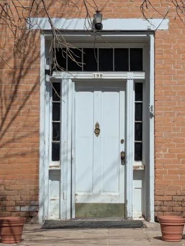 Brick facade entry with a paneled white door, transom window, sidelights, and a wall-mounted light fixture