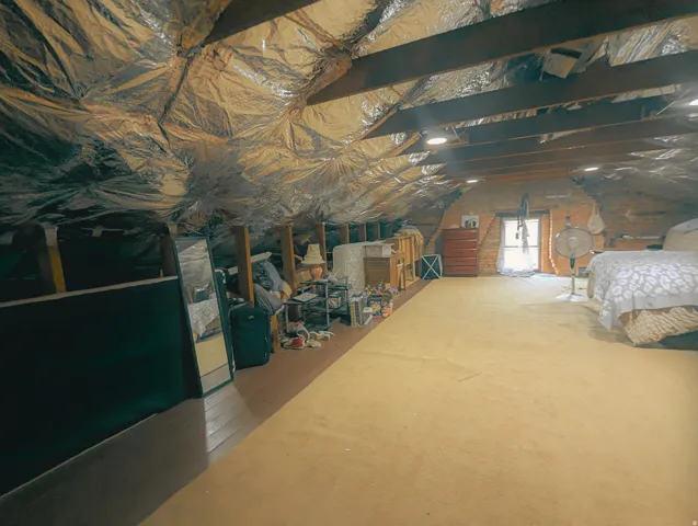Finished attic space featuring exposed wooden beams, foil-faced insulation, light gold carpeting, and built-in lighting