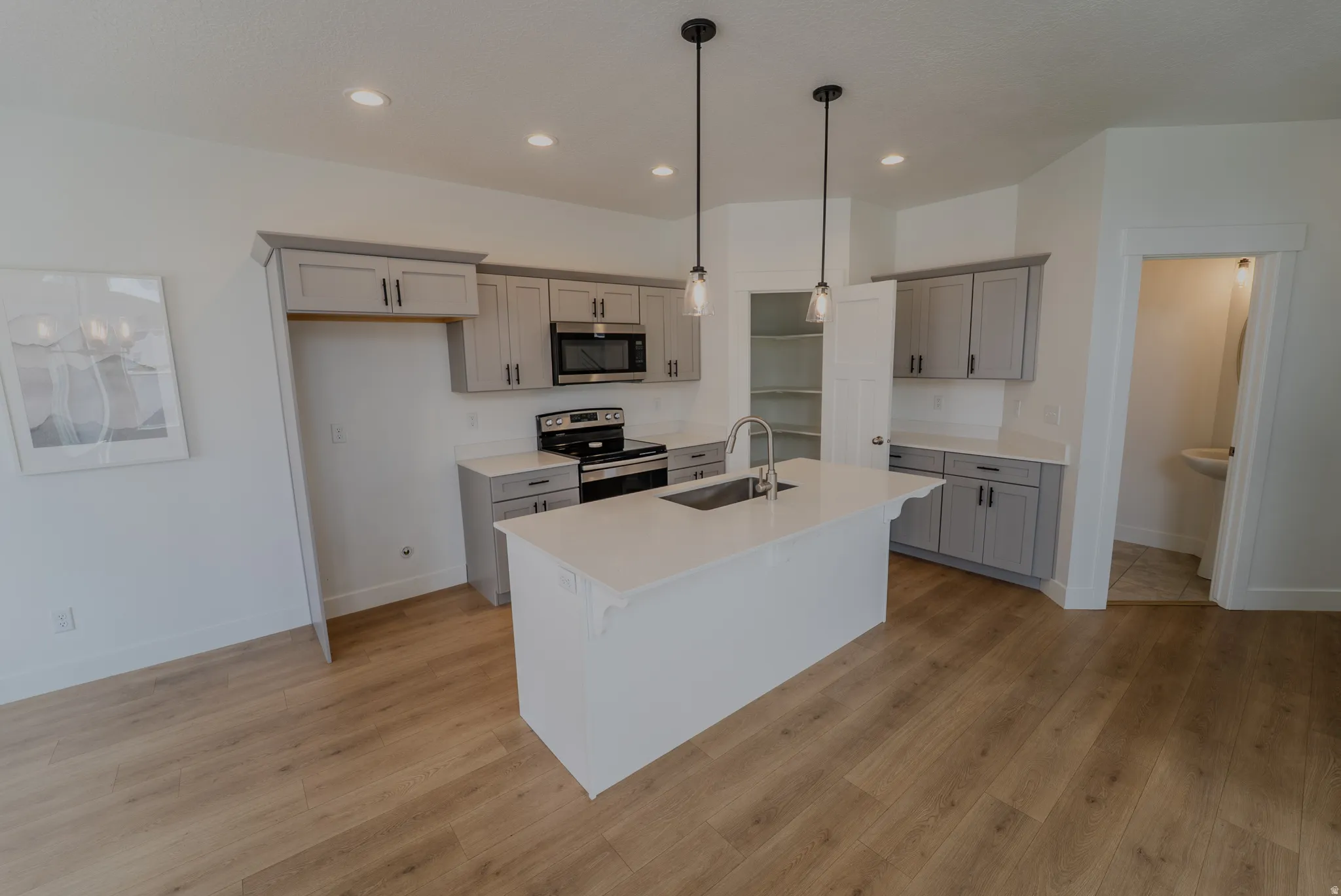 Contemporary kitchen featuring a central island with an undermount sink, pendant lighting, light gray cabinetry, stainless steel appliances, and wood-finish flooring