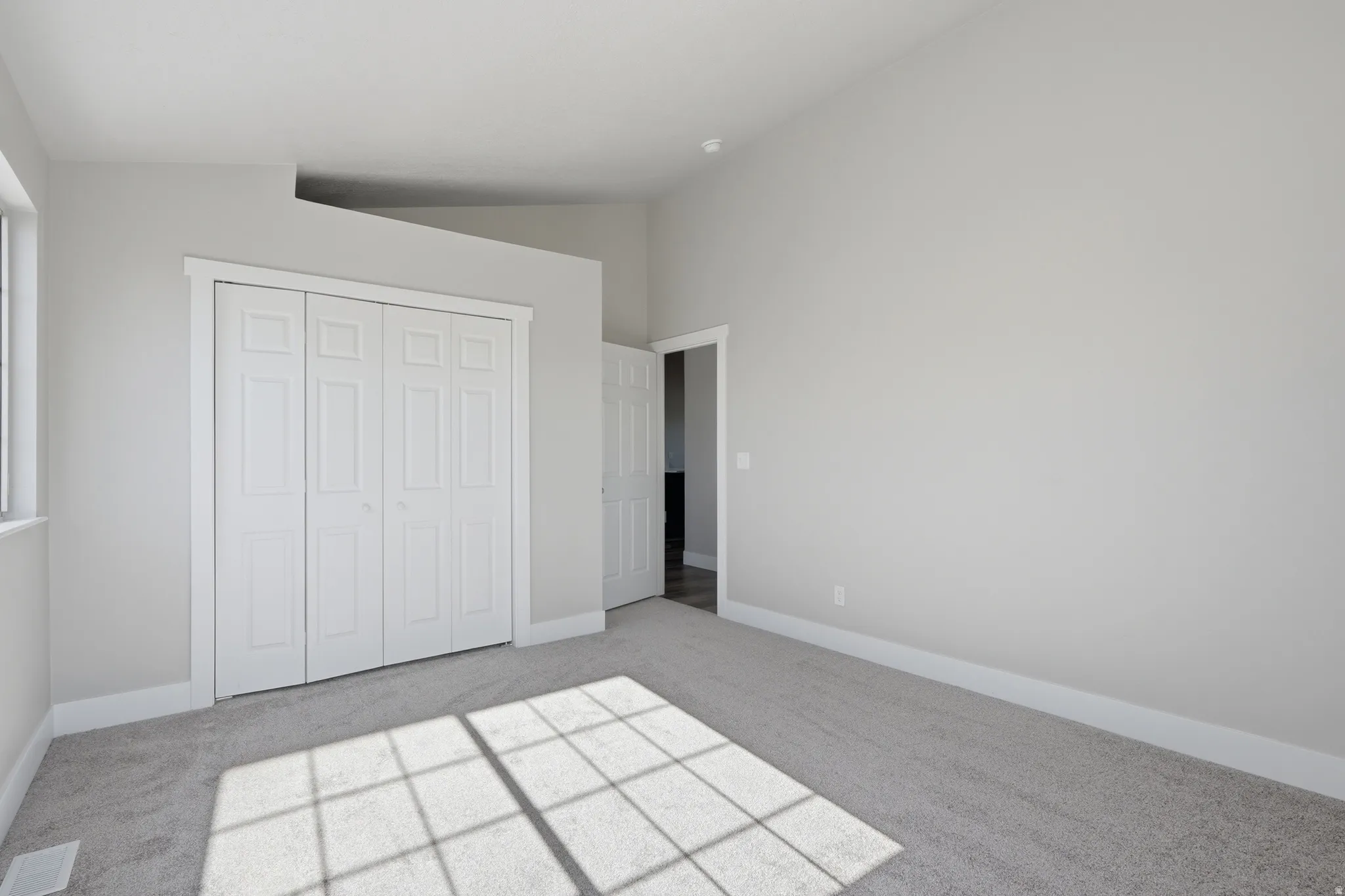 Spacious room featuring light gray wall paint, light gray carpet, white bi-fold closet doors, white baseboards, and a vaulted ceiling