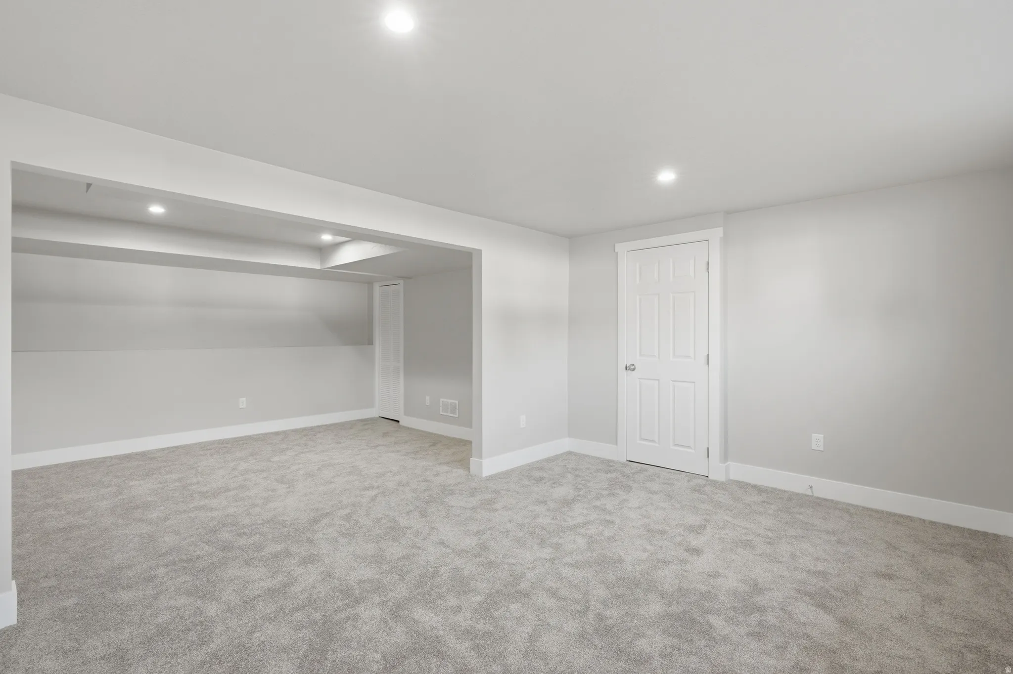 Spacious finished room featuring plush gray carpeting, recessed lighting, white baseboards, and a white paneled door