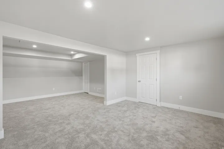Spacious finished room featuring plush gray carpeting, recessed lighting, white baseboards, and a white paneled door