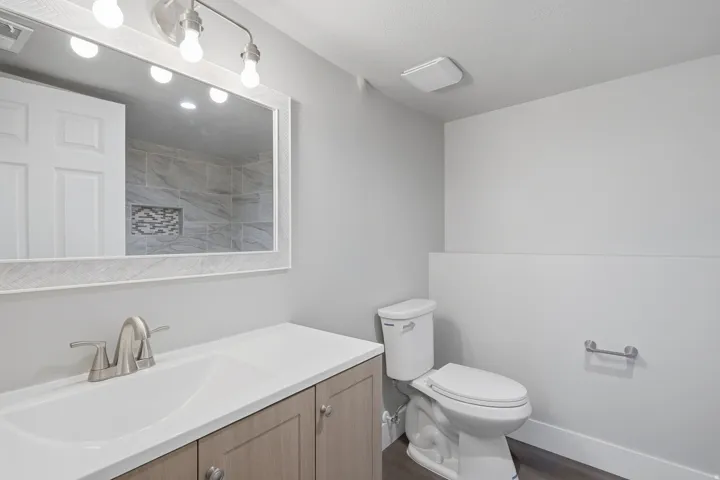 Vanity with integrated sink and brushed nickel faucet, framed mirror with vanity lighting, wood-finish cabinetry, and white toilet