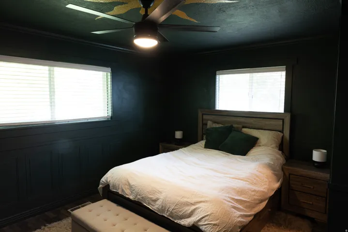 Dark-toned room featuring two windows with horizontal blinds, wall paneling, and a ceiling fan with integrated lighting