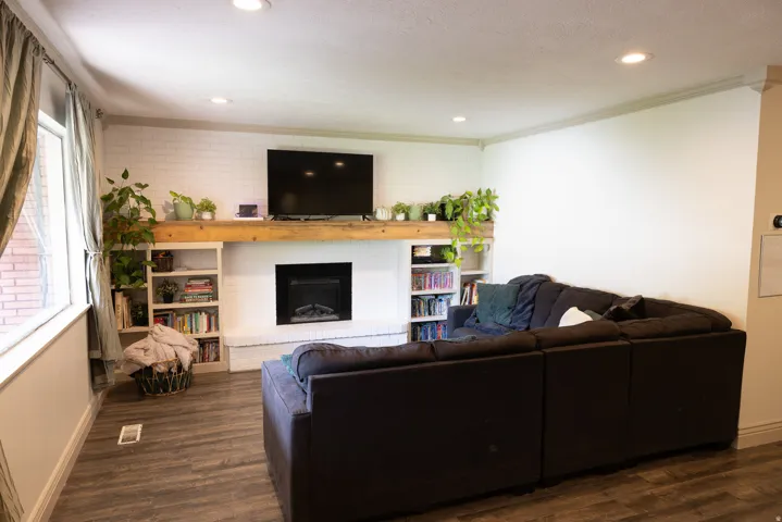 Living area featuring a white brick fireplace with a wood-beam mantel, recessed lighting, built-in shelving, and wood-finish flooring
