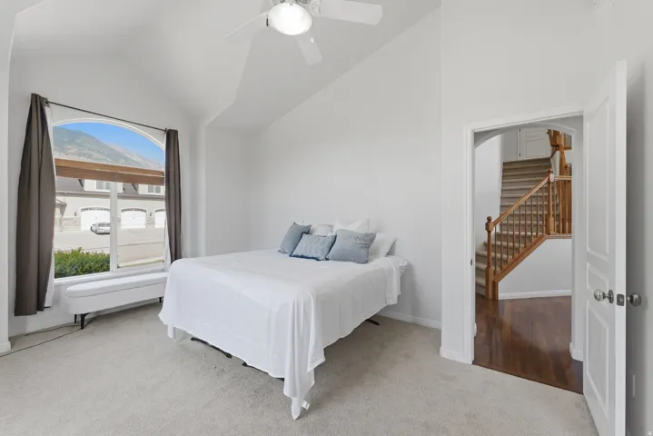 Vaulted ceiling bedroom featuring an arched window, mountain backdrop, ceiling fan with light fixture, and plush carpet flooring