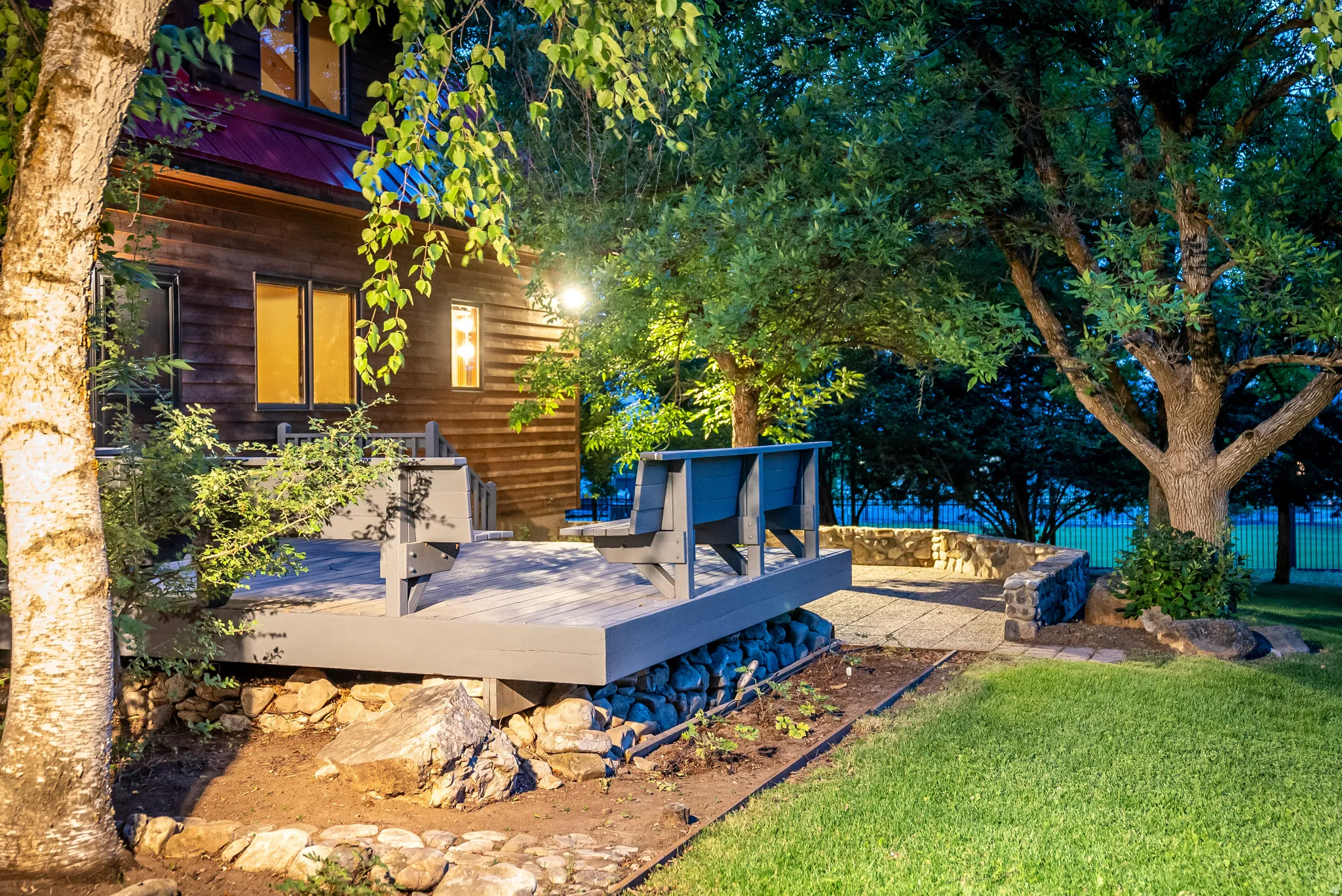 Wood-sided exterior featuring a raised deck with integrated seating, stone retaining wall, paved patio area, and mature trees