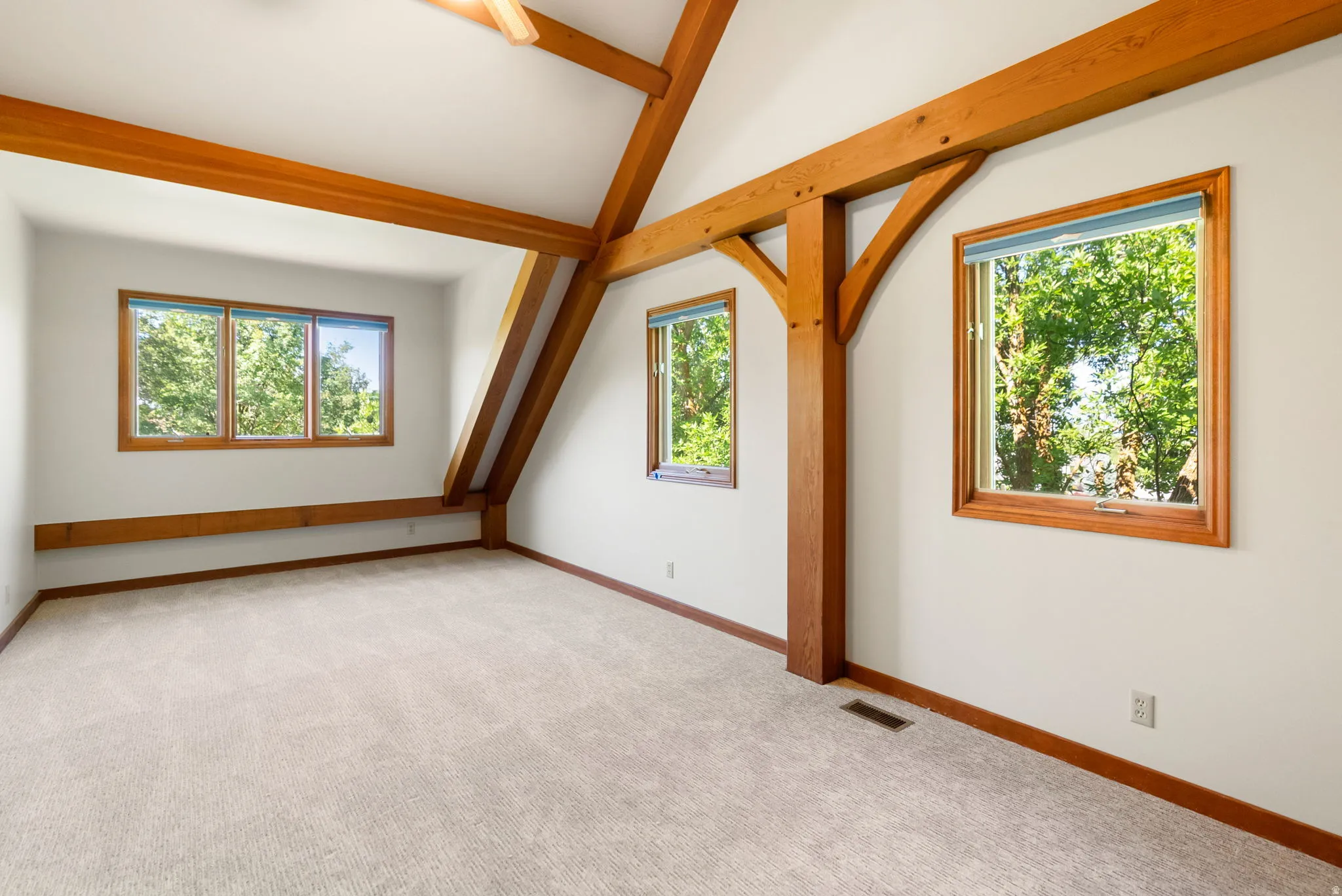 Vaulted ceiling with exposed wood beams, multiple windows, light-toned carpeting, and wood window trim