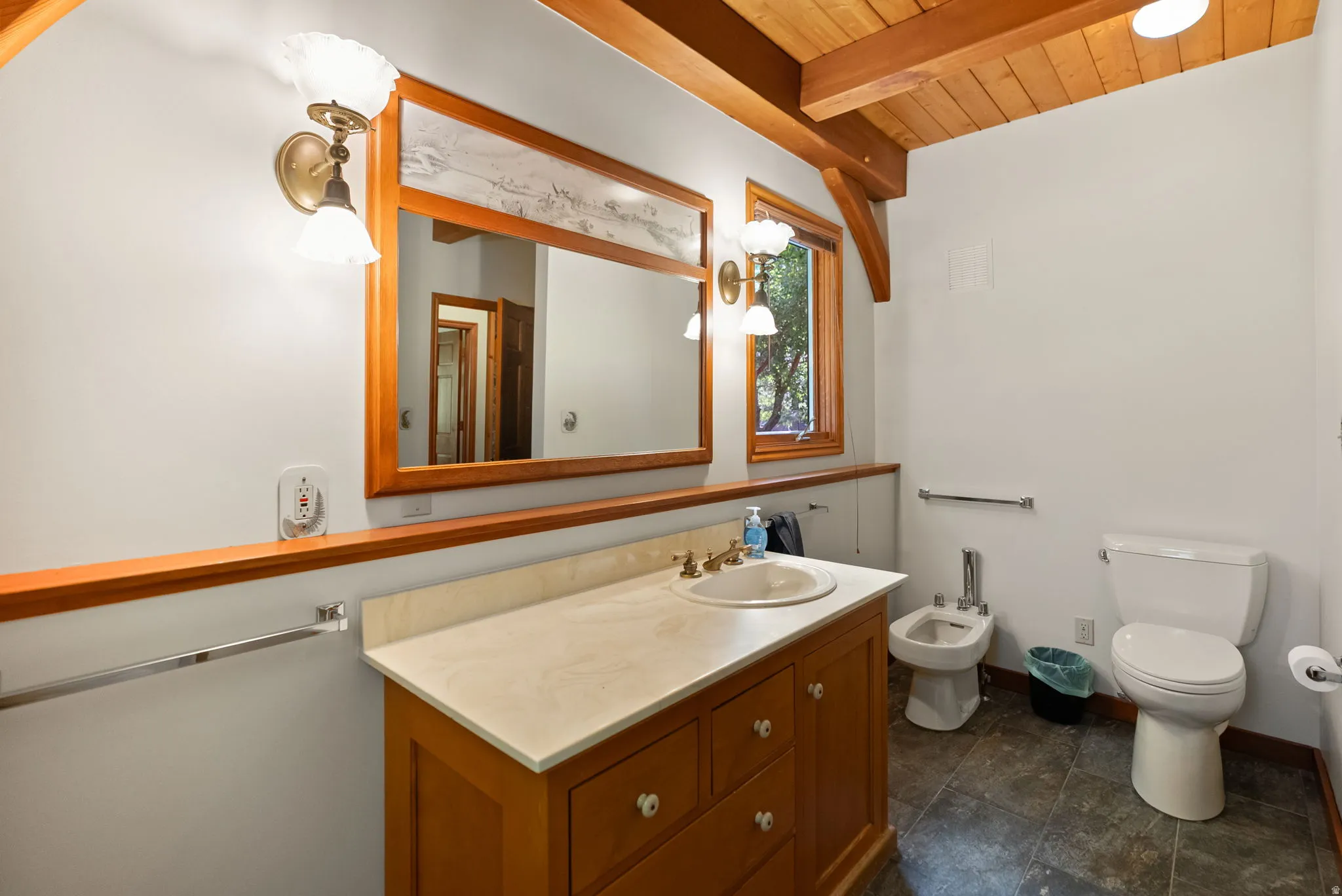 Bathroom featuring a wood-finish vanity with an integrated sink and light-toned countertop, a large wood-framed mirror, and a window with wood trim