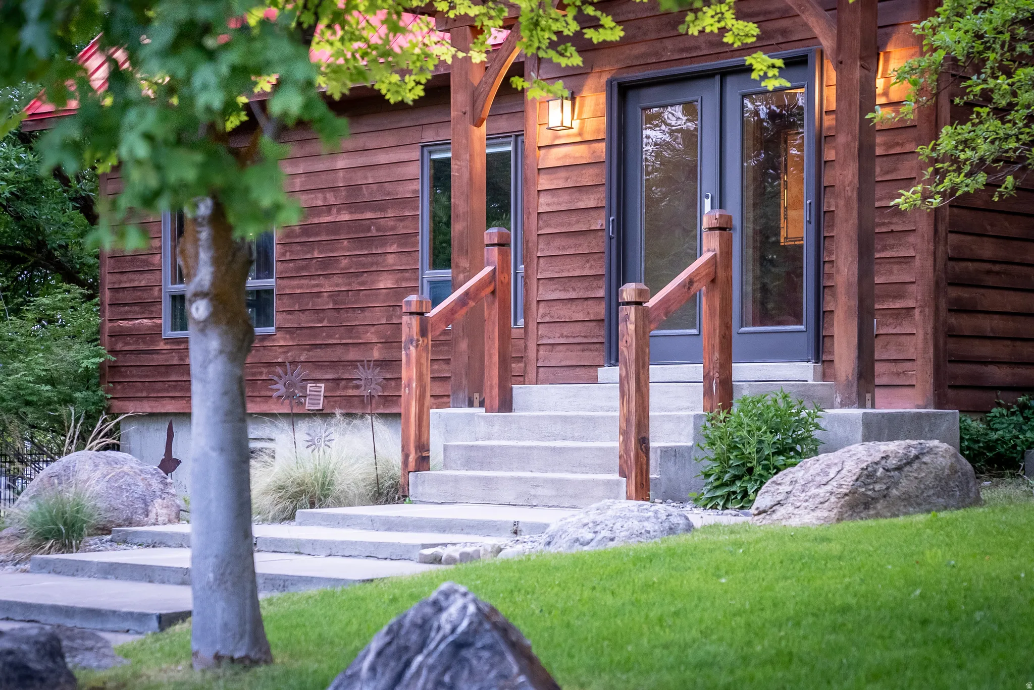 Wood-sided exterior featuring concrete steps with wood railings, a dark-framed double-door entry, exterior wall sconce, and mature landscaping