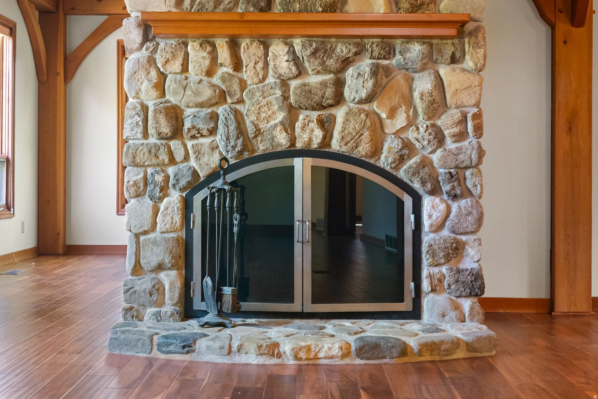 Floor-to-ceiling stone fireplace with an arched glass door enclosure, a stone hearth, a wood mantel, and wood-finish flooring
