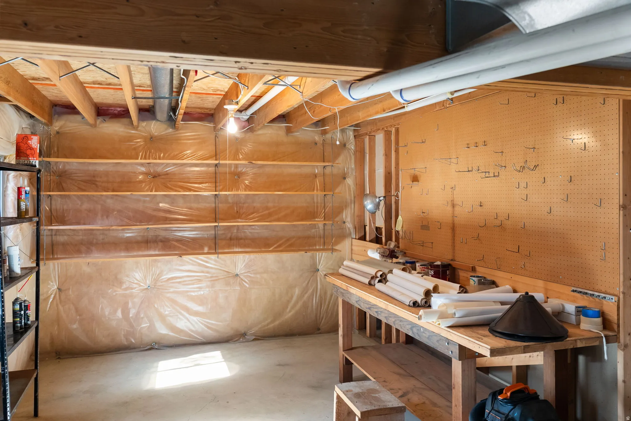 Unfinished basement featuring exposed wood beams, concrete flooring, built-in shelving, a workbench with pegboard, and visible utility lines