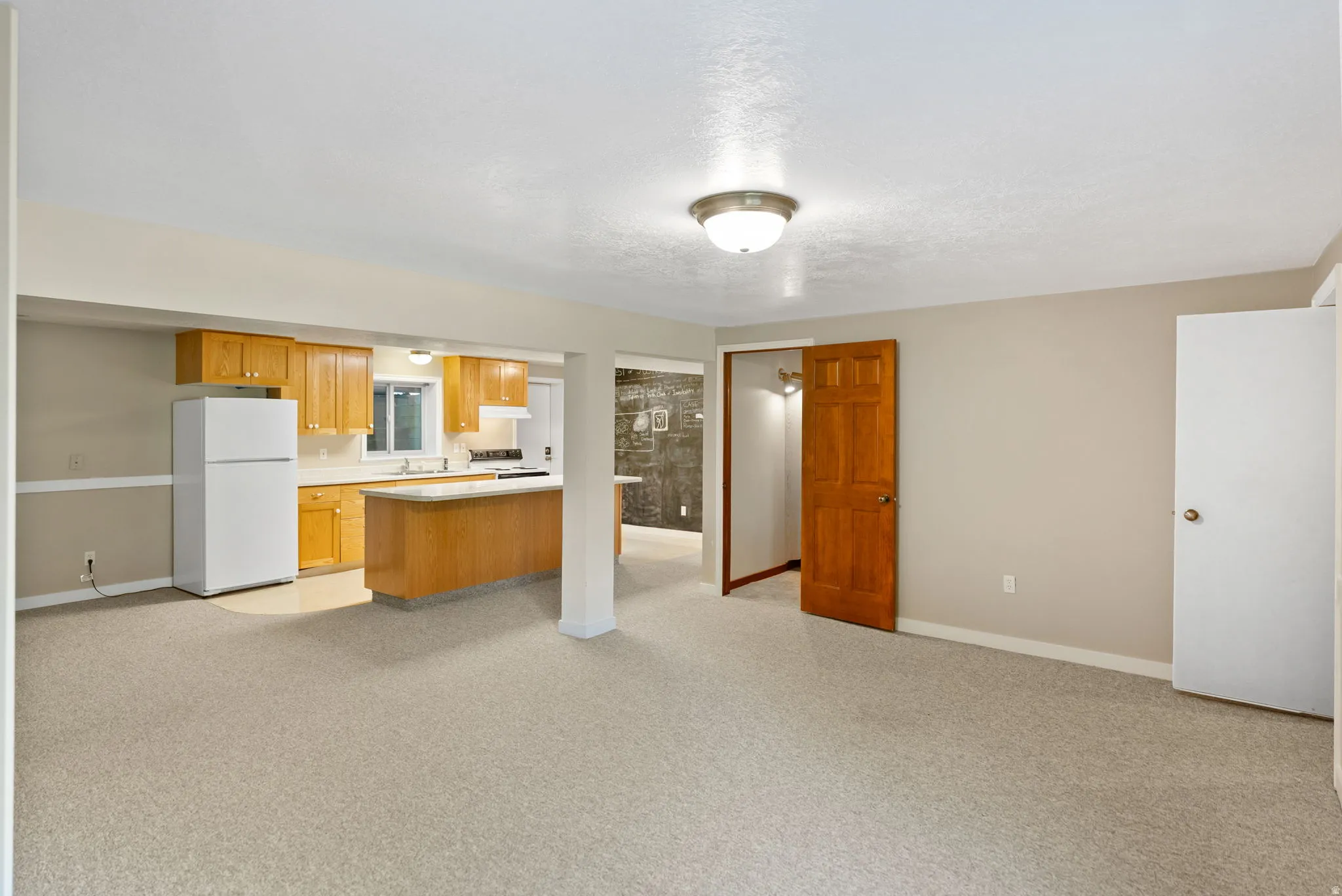 Open-concept living space featuring light-toned carpeting and a kitchen area with wood cabinetry, white appliances, and a peninsula
