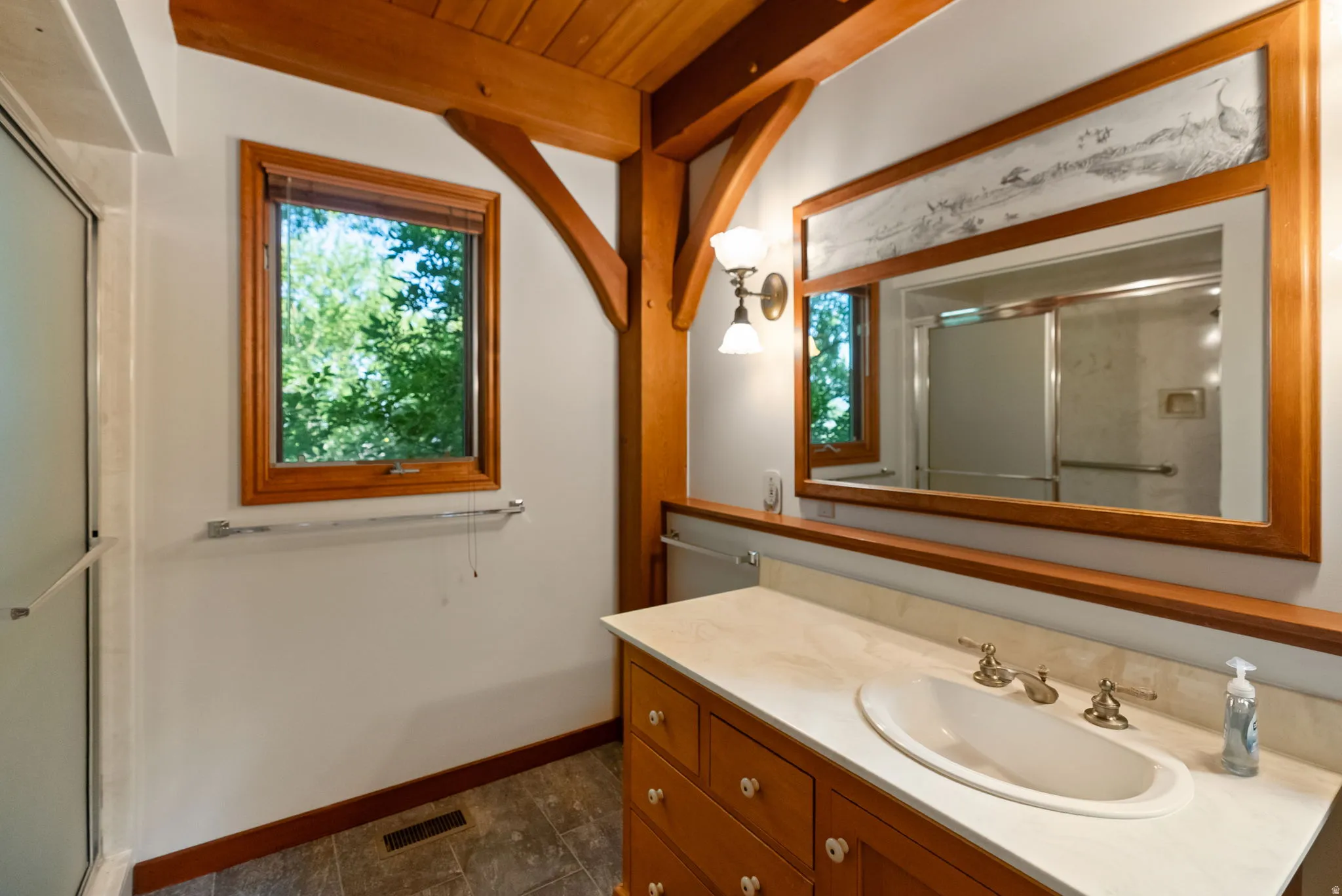 Bathroom featuring exposed timber beams, a wood-framed window, and a vanity with a light-toned countertop and wood cabinetry