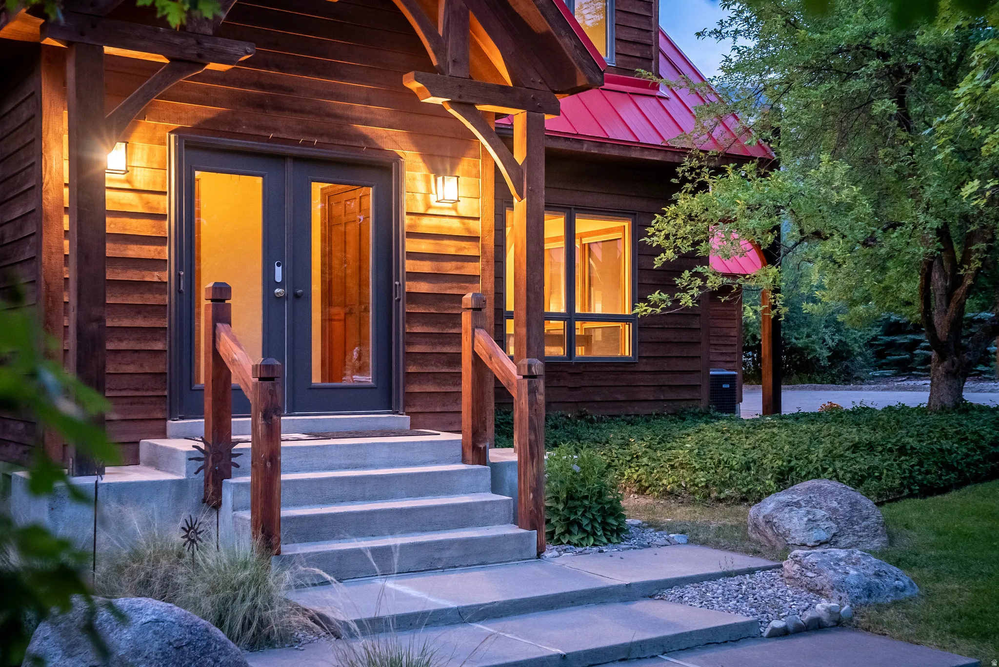 Timber-frame entrance featuring double glass doors, a red metal roof, concrete steps, wood railings, and exterior wall sconces