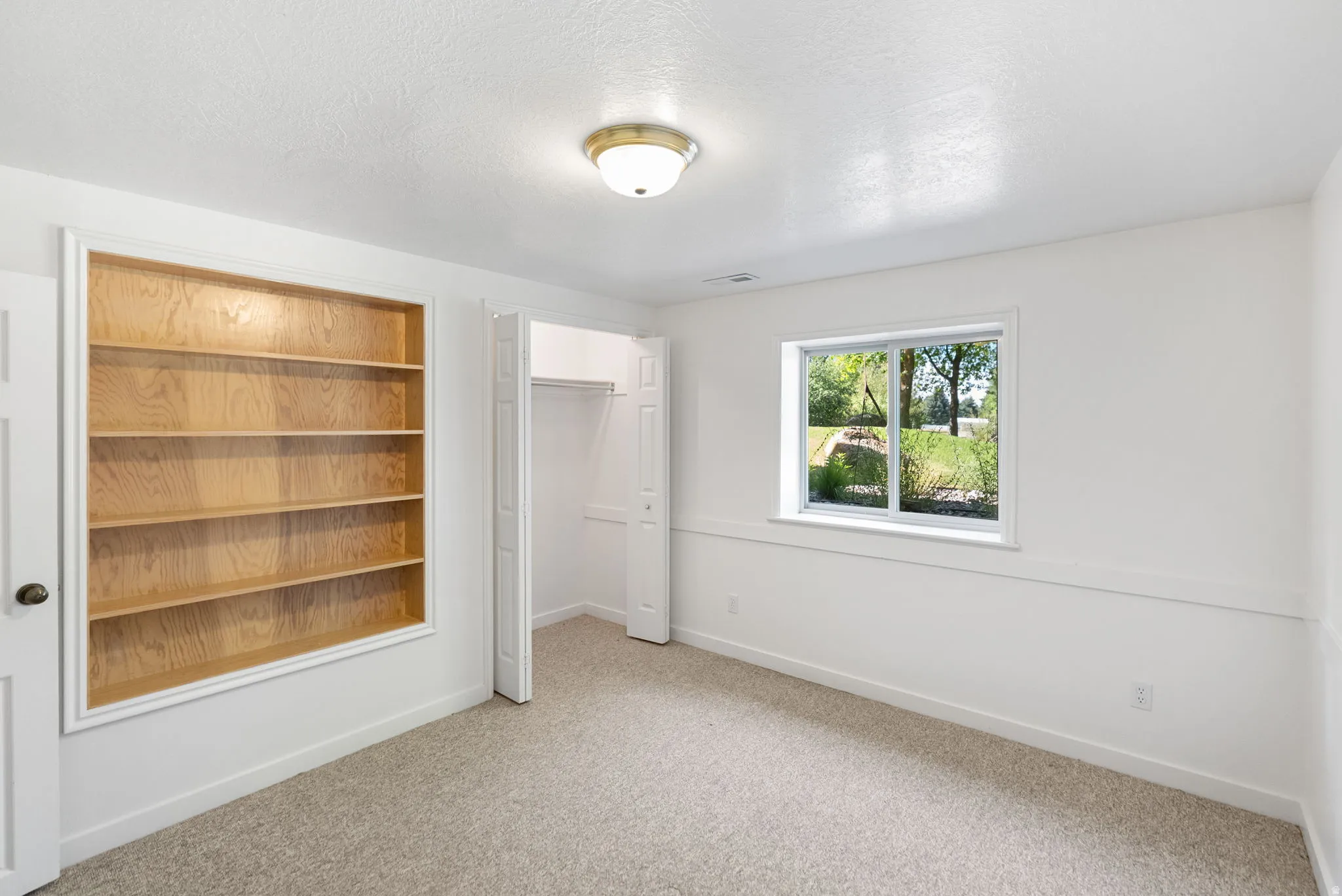 Carpeted room featuring a built-in wood-finish shelving unit, a window, a bi-fold closet, and a ceiling-mounted light fixture
