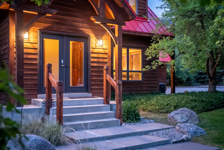 Timber-frame entrance featuring double glass doors, a red metal roof, concrete steps, wood railings, and exterior wall sconces