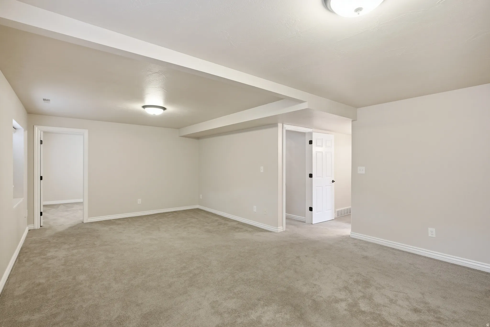 Spacious room featuring neutral carpeting, light-colored walls, recessed ceiling beams, and overhead flush-mount lighting fixtures