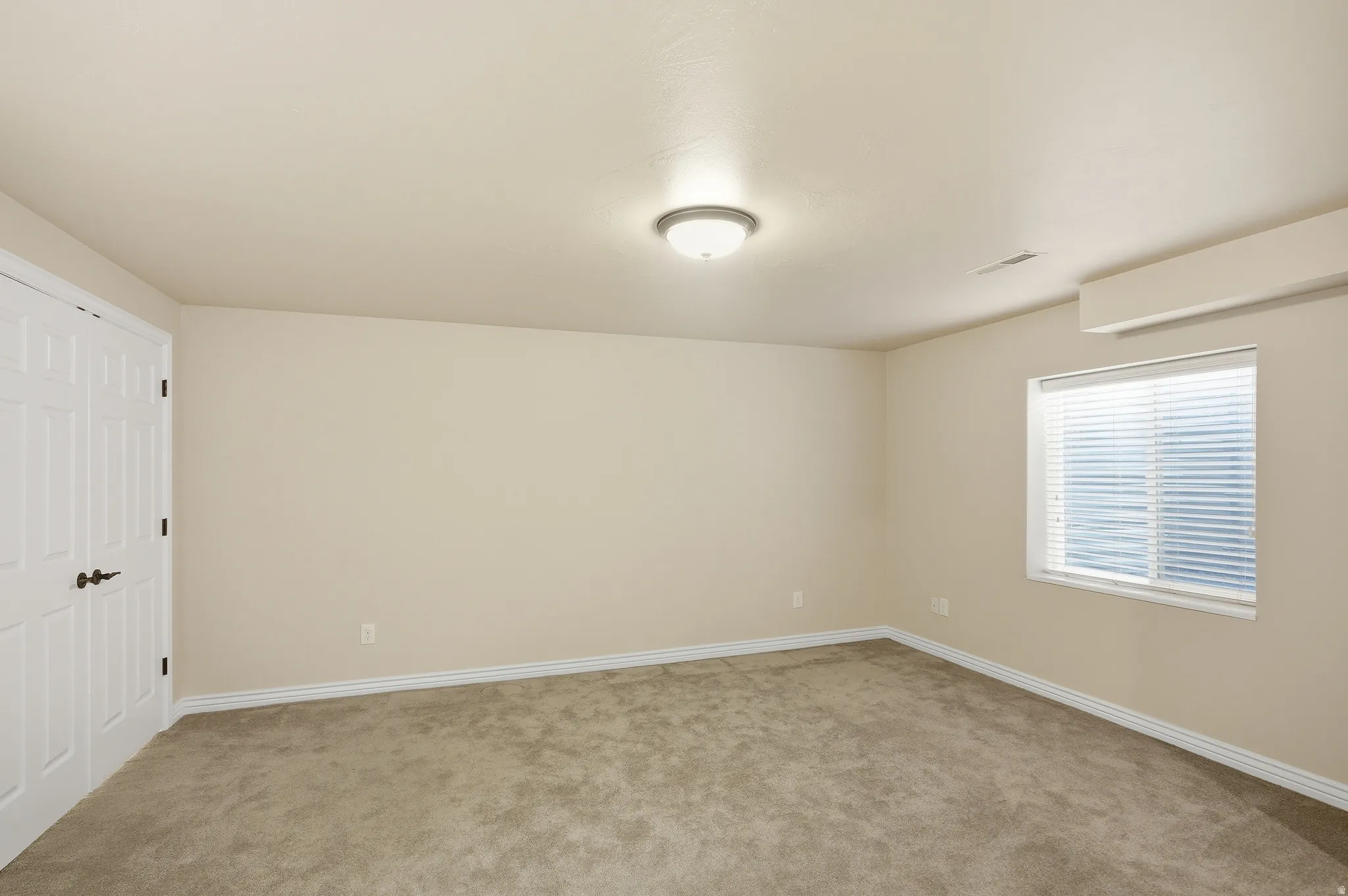 Spacious room featuring neutral wall paint, light brown carpeting, a window with horizontal blinds, and a built-in wall-mounted unit