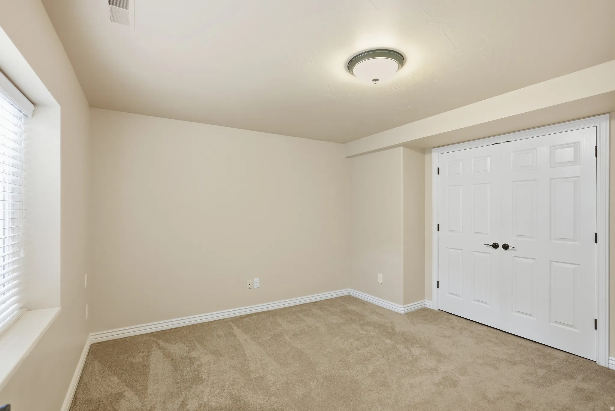 Neutral-toned room featuring a window with blinds, light tan carpeting, and a flush-mount ceiling light