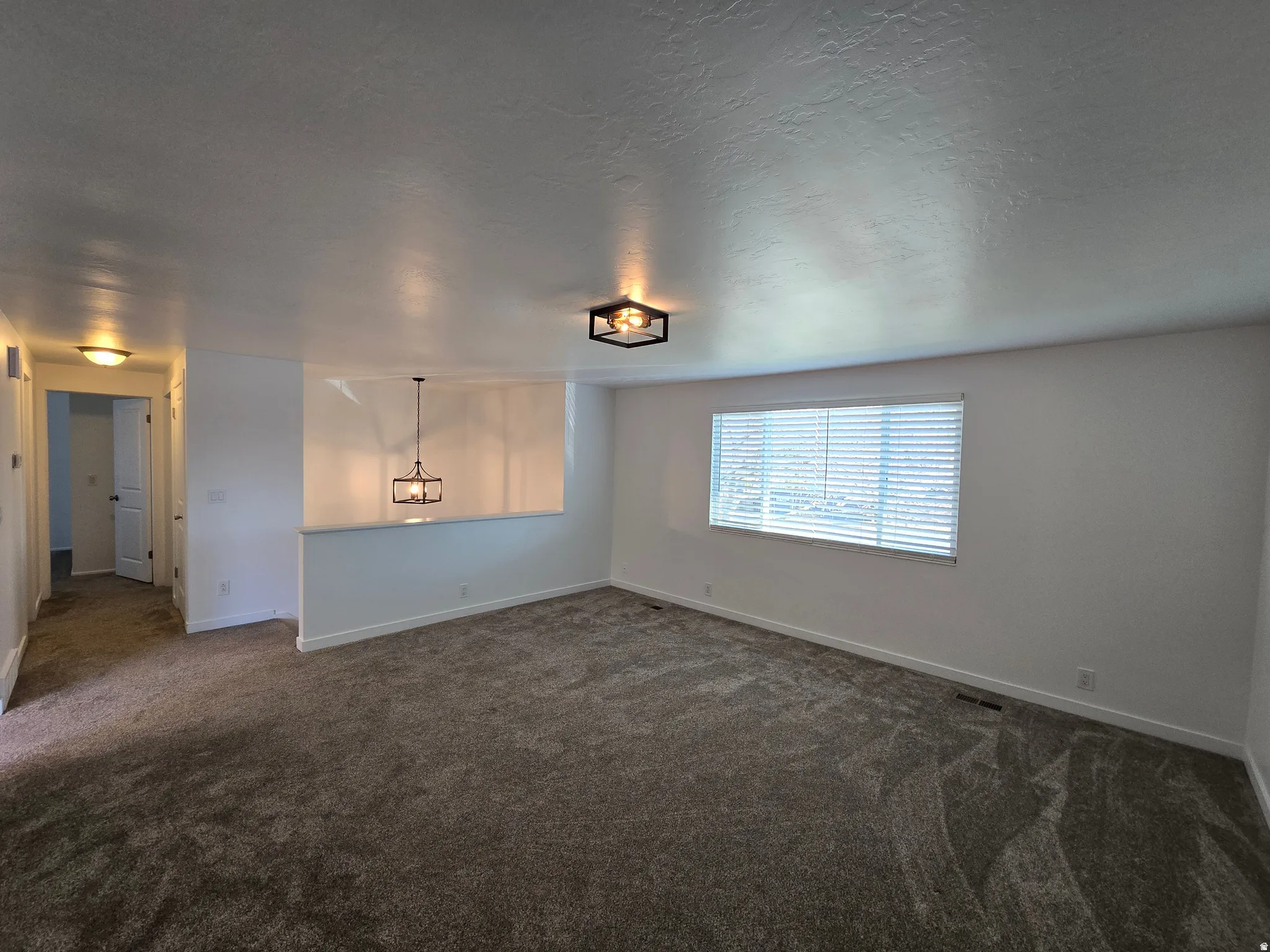 Spacious interior room featuring plush gray carpeting, white painted walls, and a large window with horizontal blinds