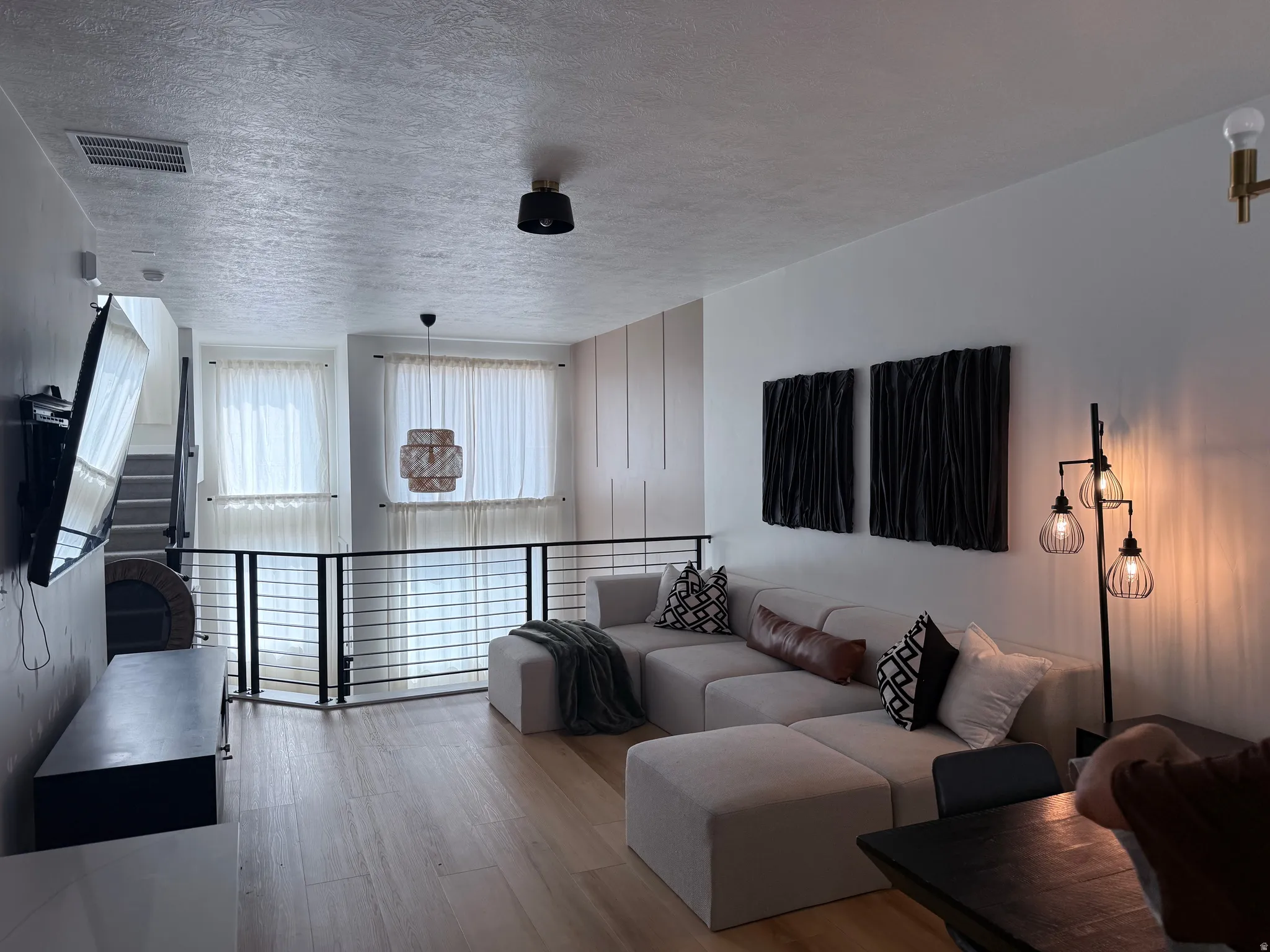 Open-concept living area with wood-finish flooring, a contemporary railing system, and multiple windows with sheer window treatments