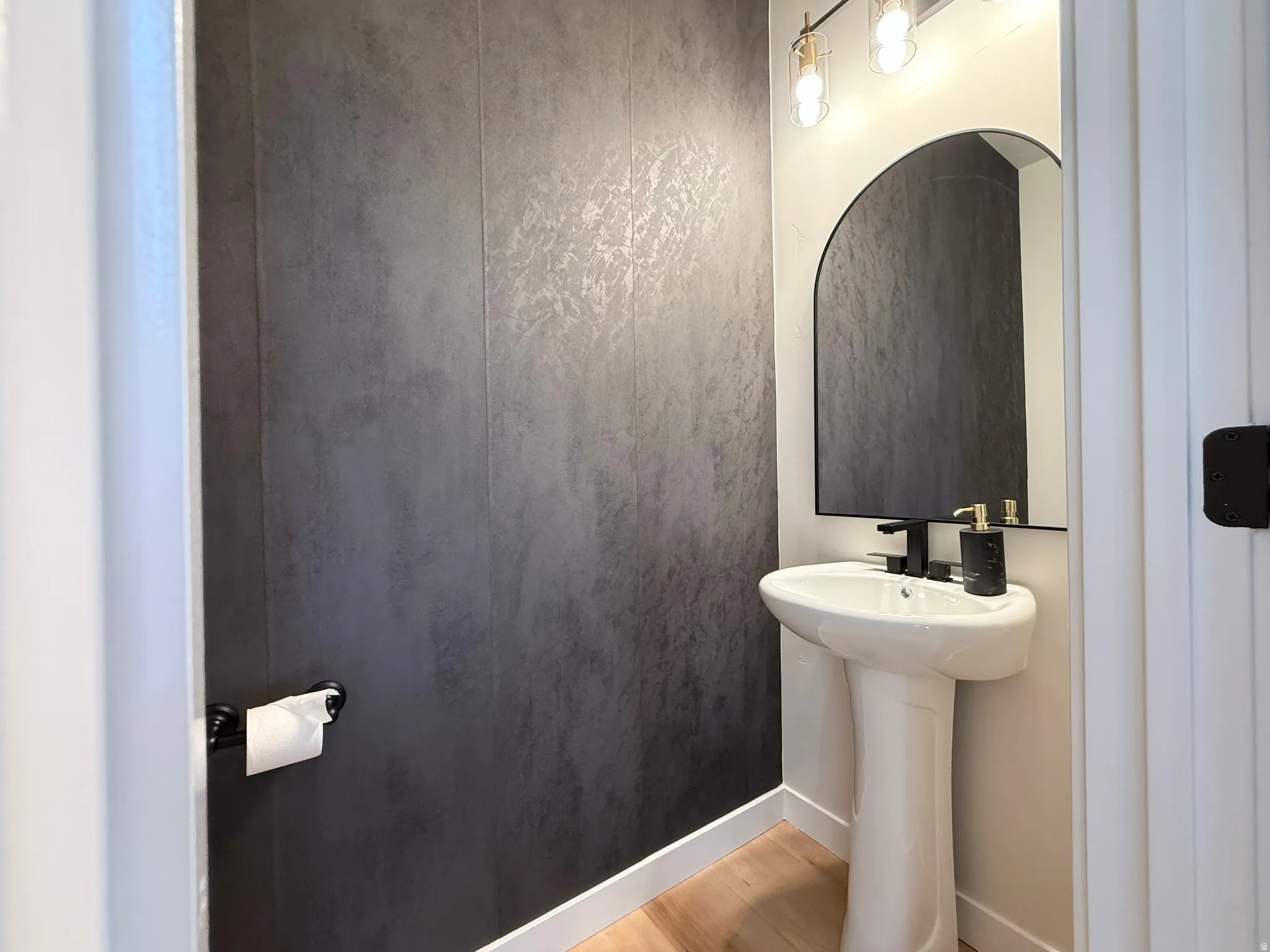 Pedestal sink with matte black faucet, arched mirror, contemporary vanity light fixture, and wood-finish flooring