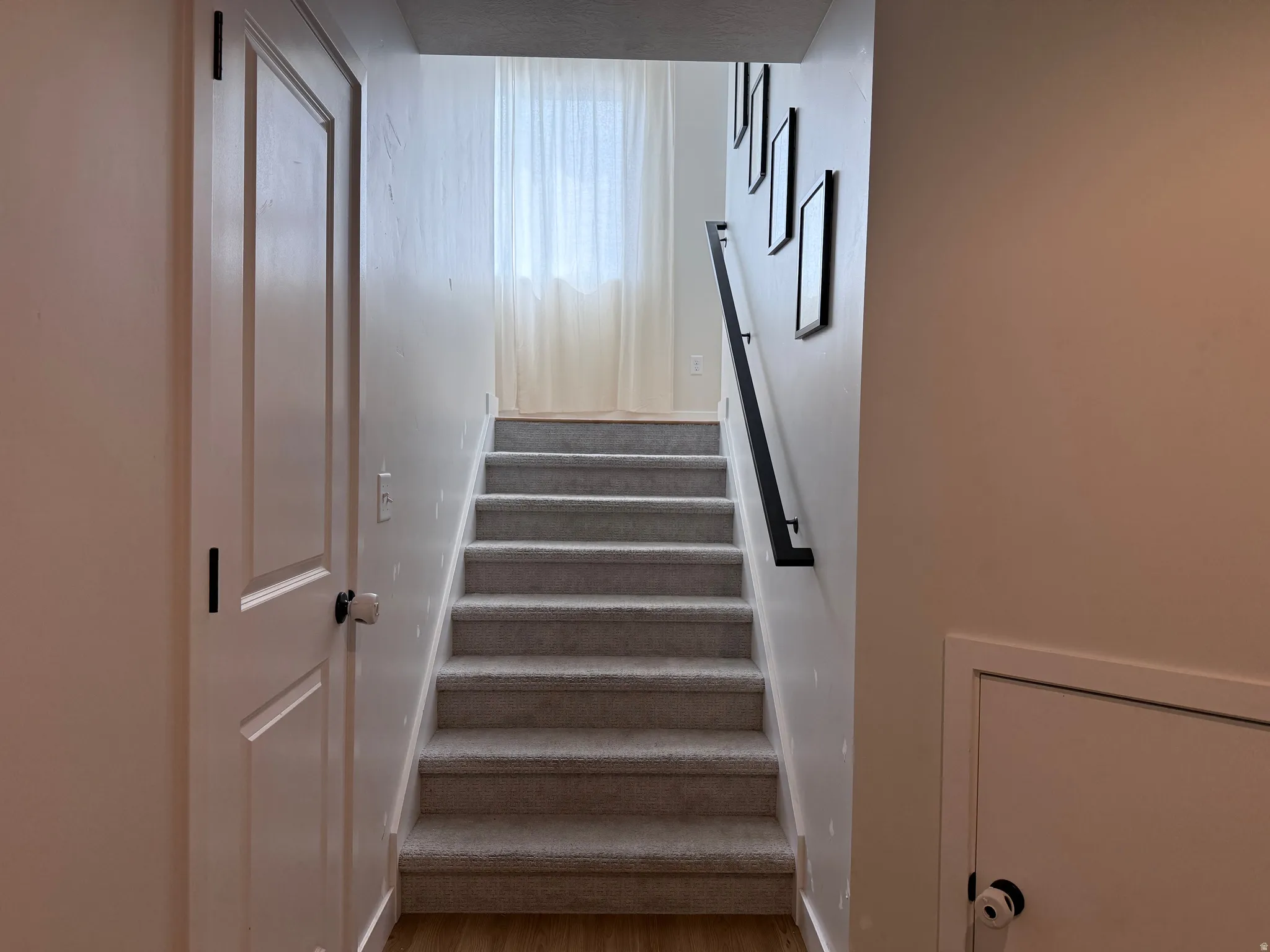 Carpeted staircase with a modern black handrail, light wood-finish landing, and a window with sheer curtains