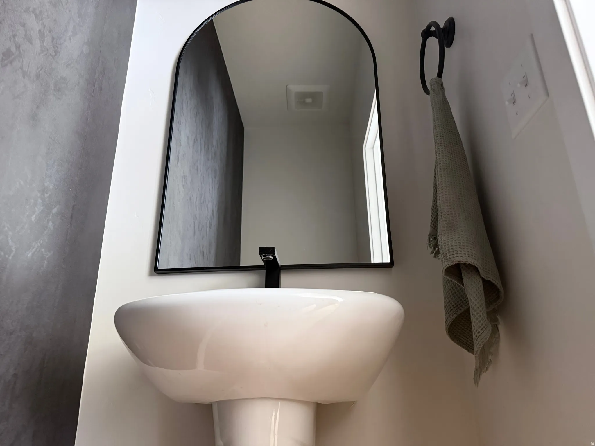 Pedestal sink with a matte black wall-mounted faucet, complemented by an arched black-framed mirror