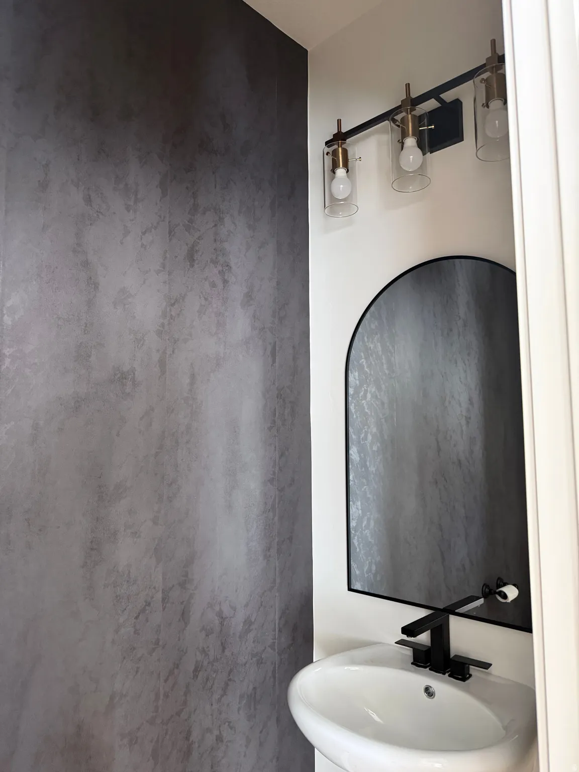 Modern powder room featuring a textured accent wall, black arched mirror, and a contemporary vanity light fixture with clear glass shades
