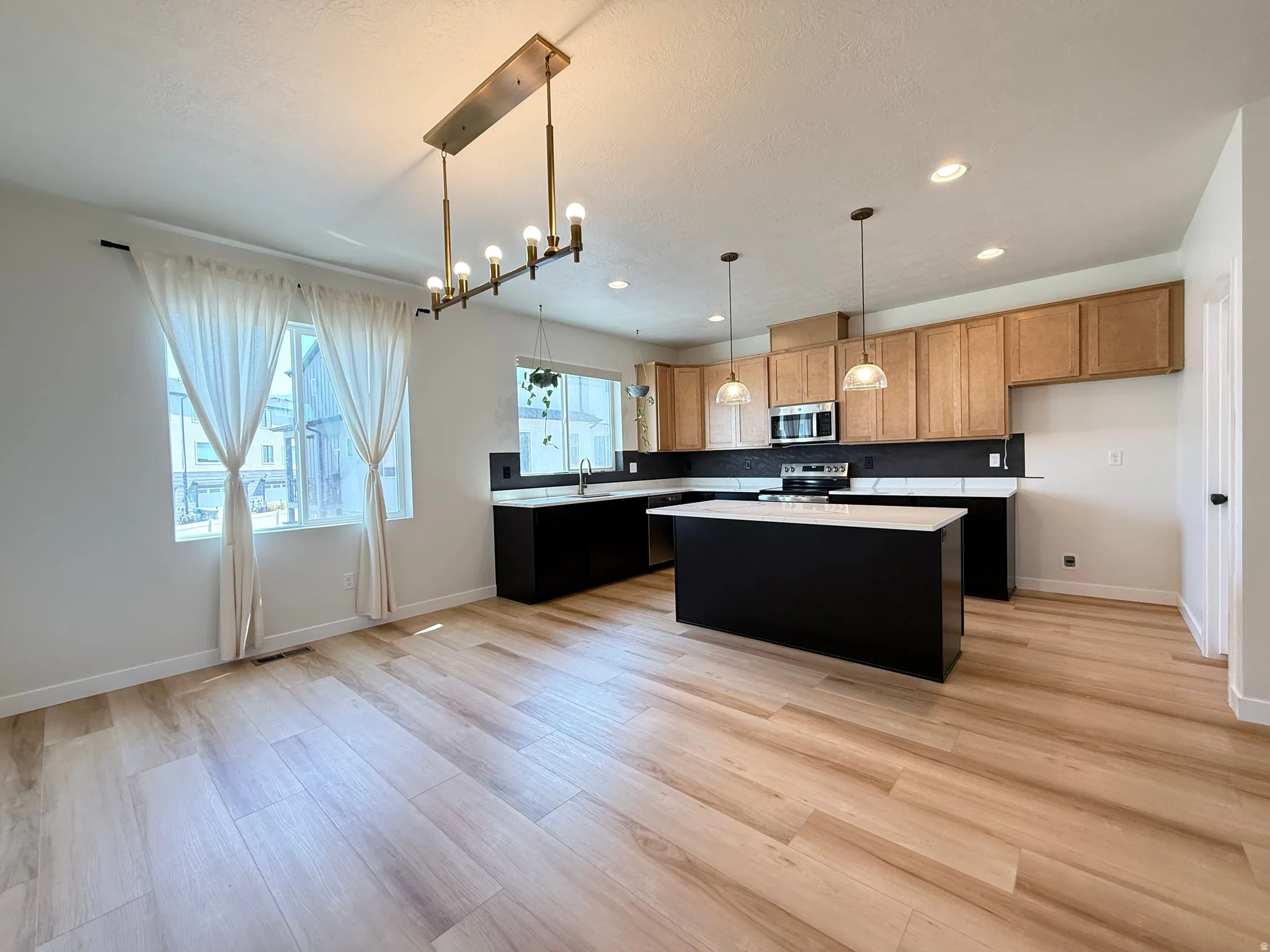 Open-concept kitchen and dining area featuring wood-finish flooring, a linear chandelier, a kitchen island with pendant lighting, and stainless steel appliances