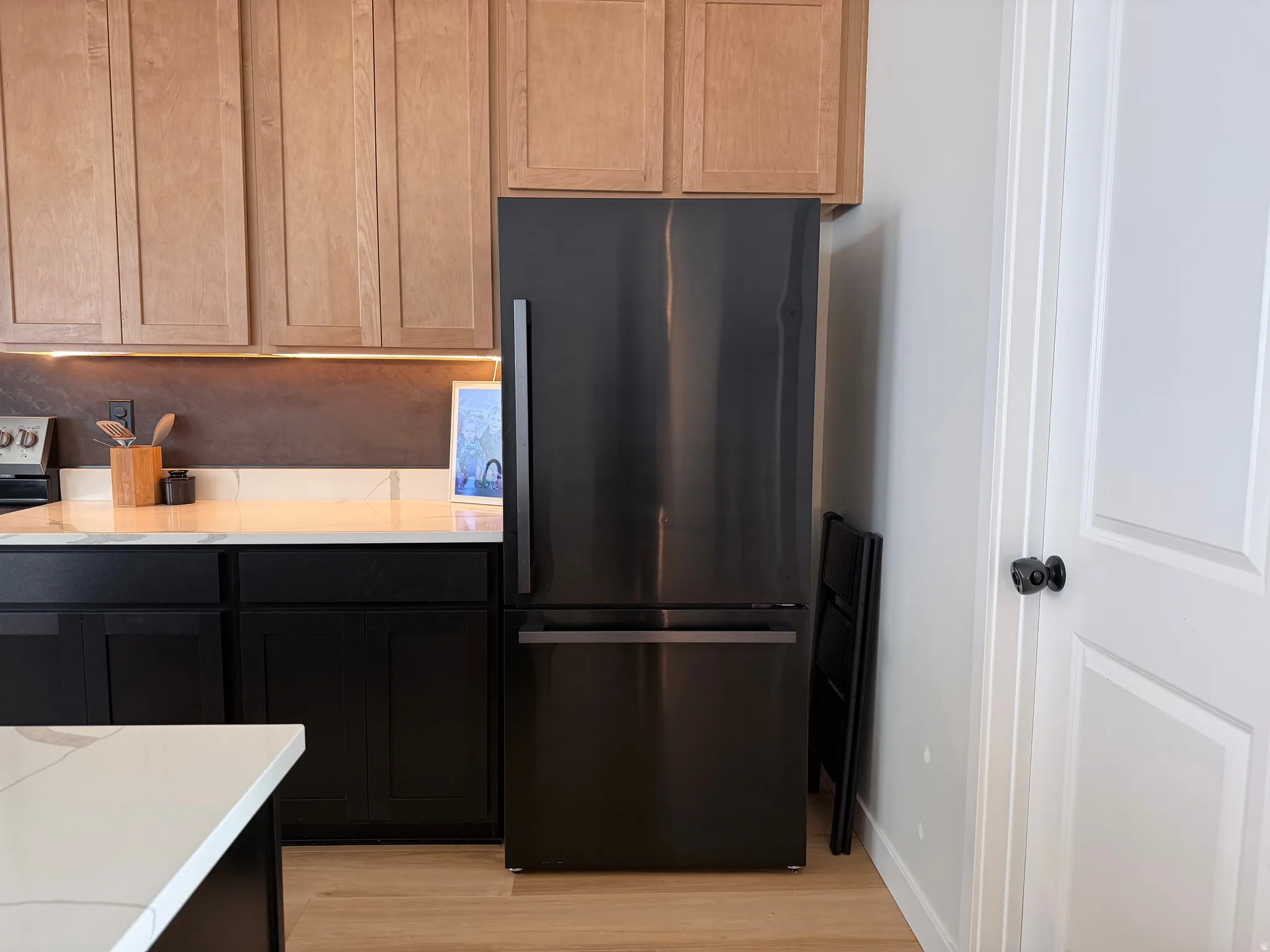 Stainless steel refrigerator integrated within a kitchen featuring light wood-finish upper cabinetry, black lower cabinetry, and a white countertop with subtle veining