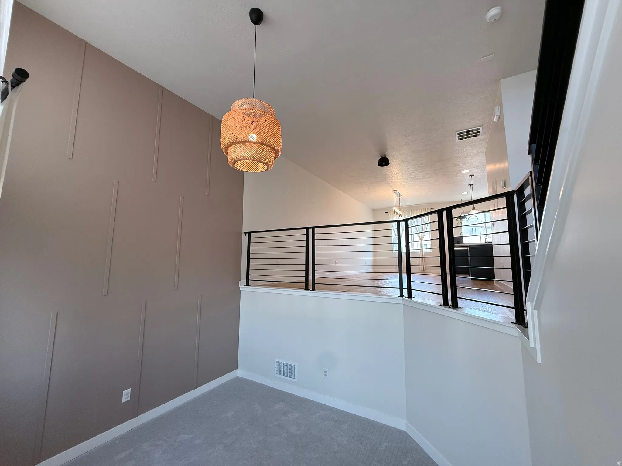 Textured accent wall, vaulted ceiling, woven pendant light fixture, black metal railing, and light wood-finish flooring