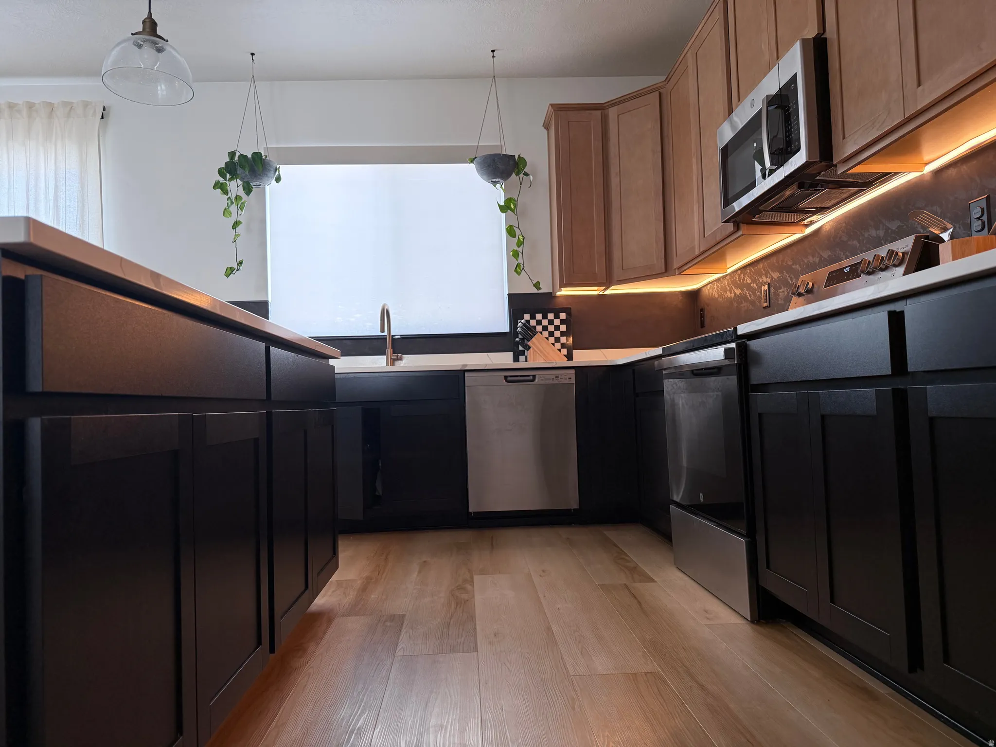 Kitchen featuring wood-finish flooring, two-tone cabinetry with dark lower cabinets and light upper cabinets, stainless steel appliances, a window above the sink, and under-cabinet lighting