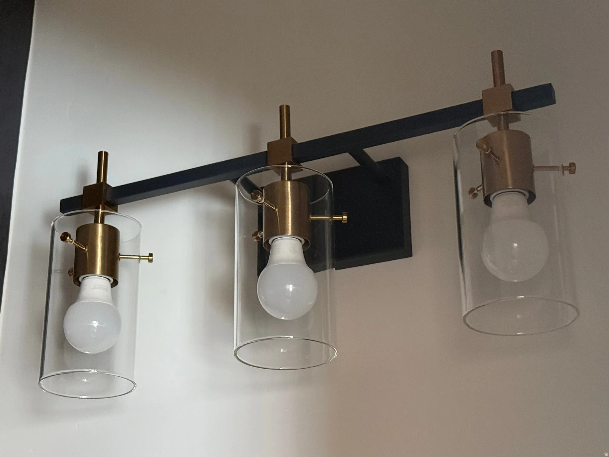 Three-light vanity fixture featuring clear glass shades and brass-finish hardware mounted on a matte black bar