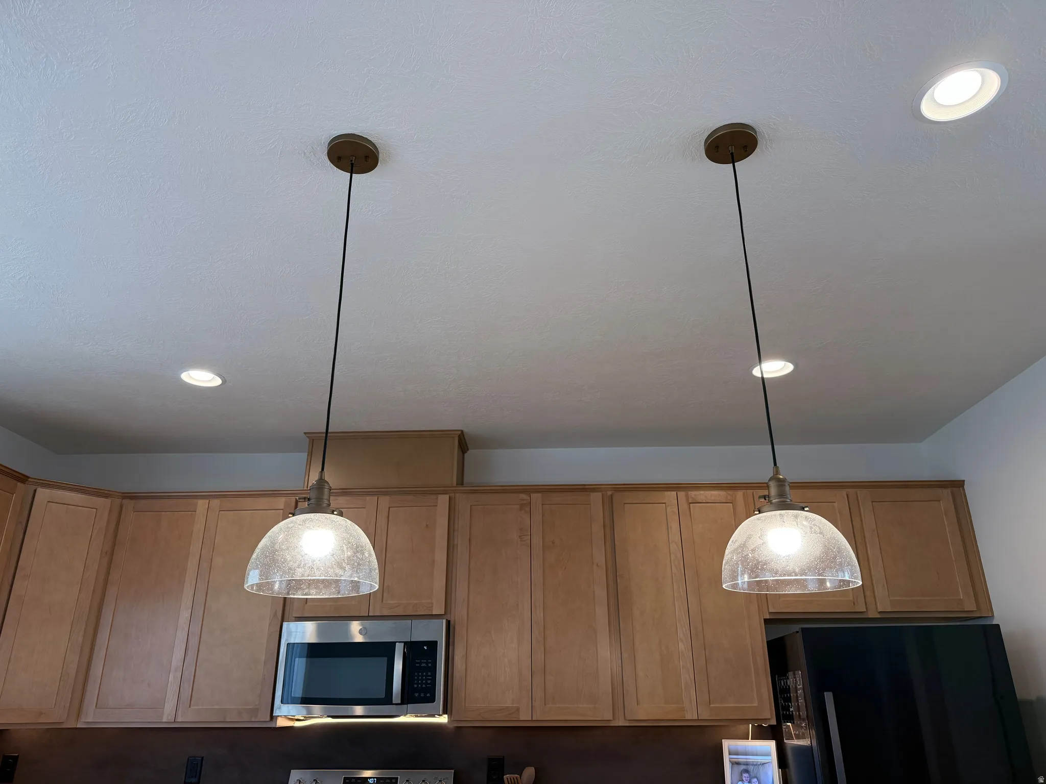Kitchen featuring two suspended pendant lights with seeded glass shades, recessed ceiling lighting, light wood-finish cabinetry, and stainless steel appliances including a microwave