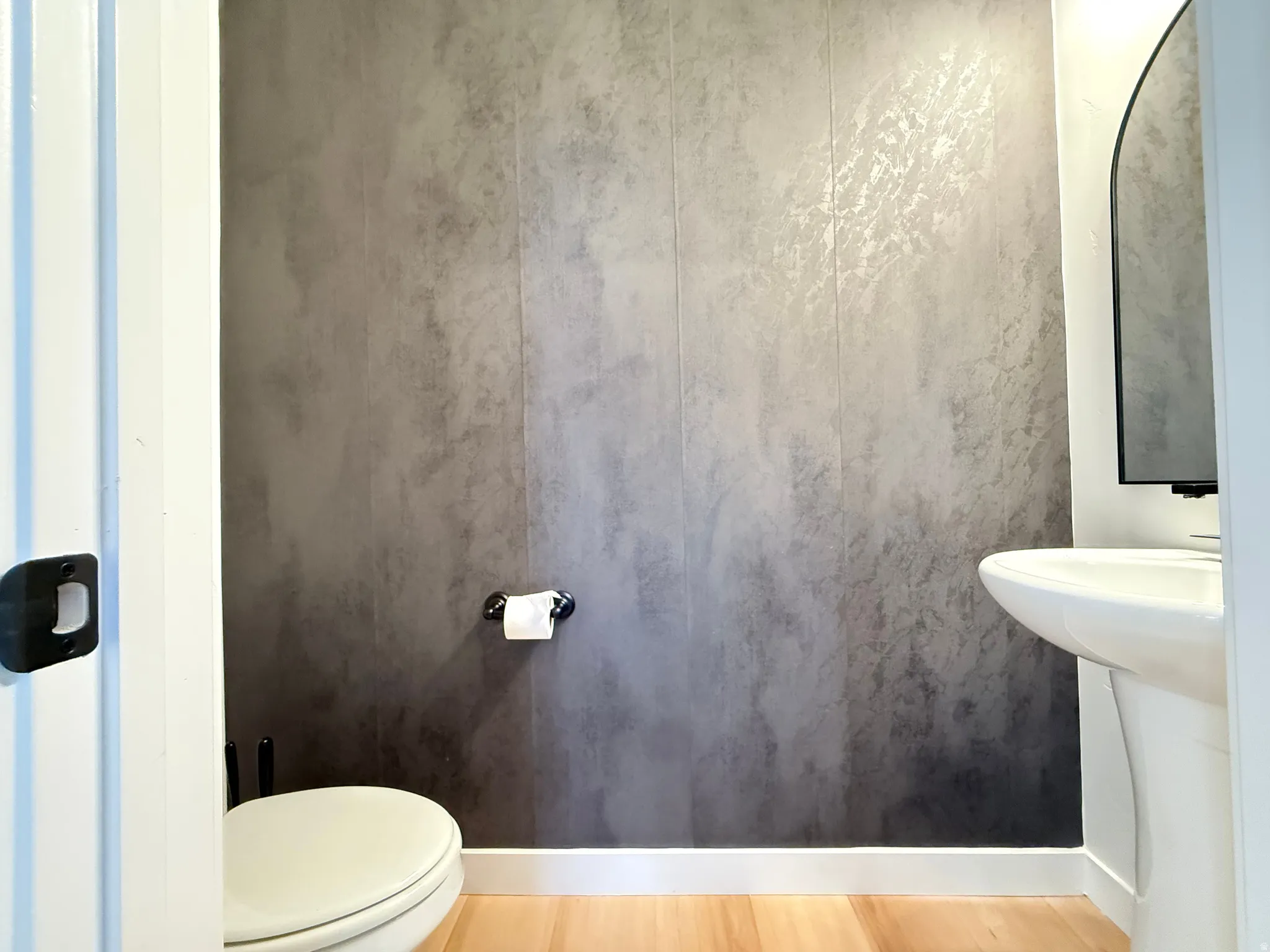 Pedestal sink with an arched mirror, textured wall covering, wood-finish flooring, and white trim