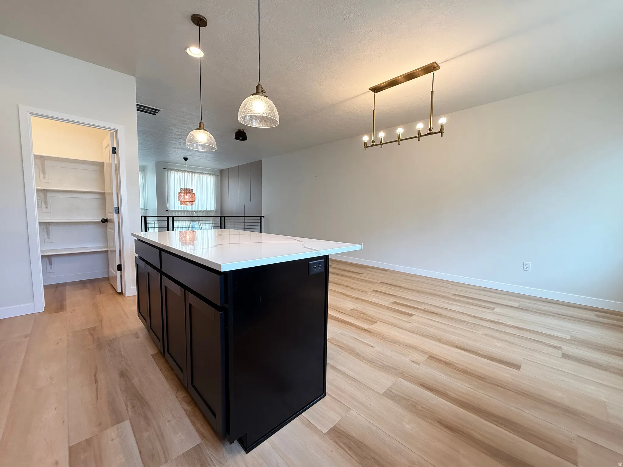 Spacious kitchen island with a white countertop and dark cabinetry, wood-finish flooring, two pendant lights, a linear chandelier, and a built-in pantry with shelving