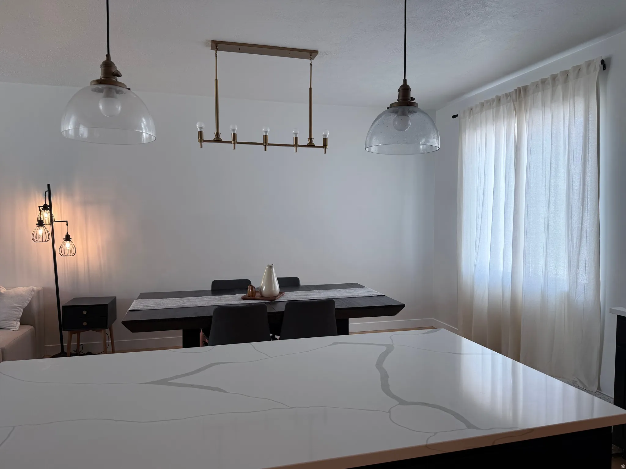 Spacious room featuring a large dining table with seating, two clear glass pendant lights, a linear chandelier, and a prominent window with sheer curtains