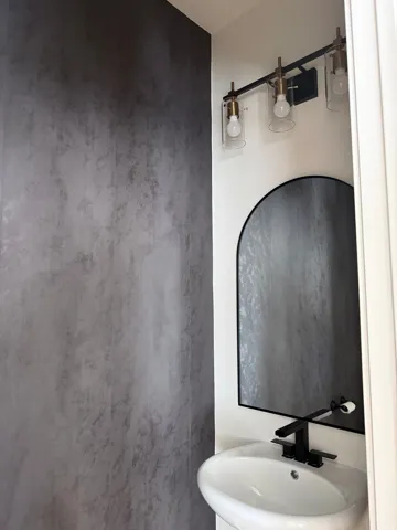 Modern powder room featuring a textured accent wall, black arched mirror, and a contemporary vanity light fixture with clear glass shades