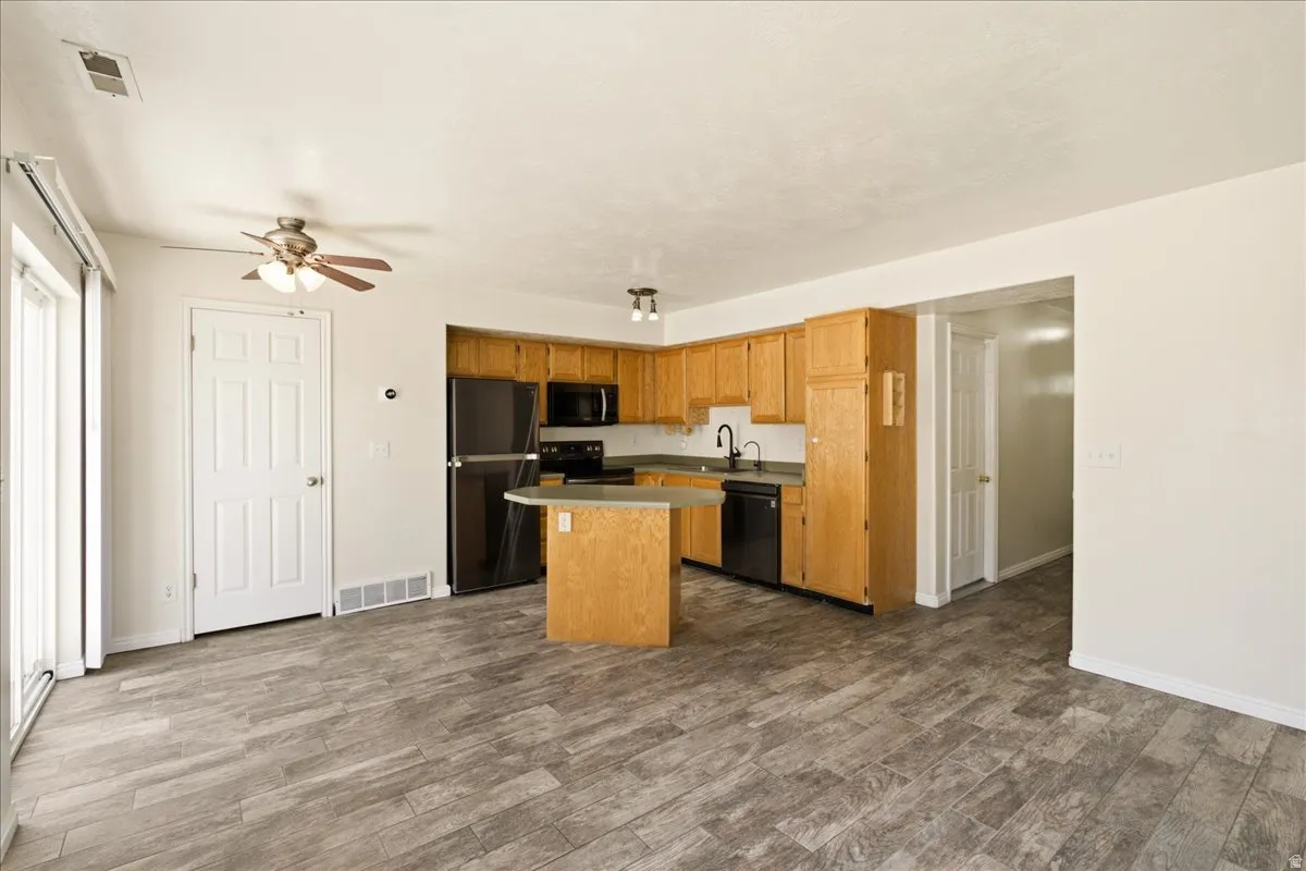 Open-concept kitchen and living area featuring wood-finish flooring, light wood cabinetry, black appliances, a central kitchen island, and sliding glass door access