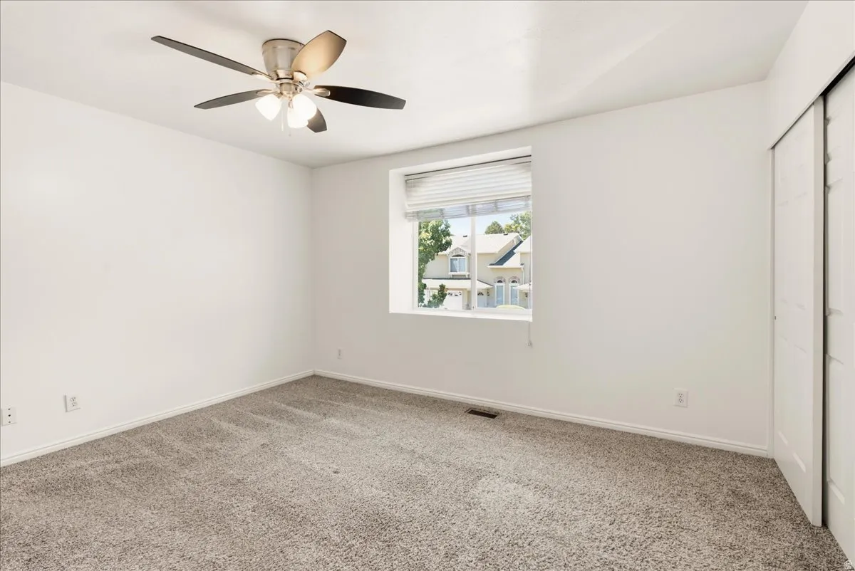 Neutral-toned carpeted interior space featuring a ceiling fan with integrated lighting, a single window with blinds, and white bi-fold closet doors